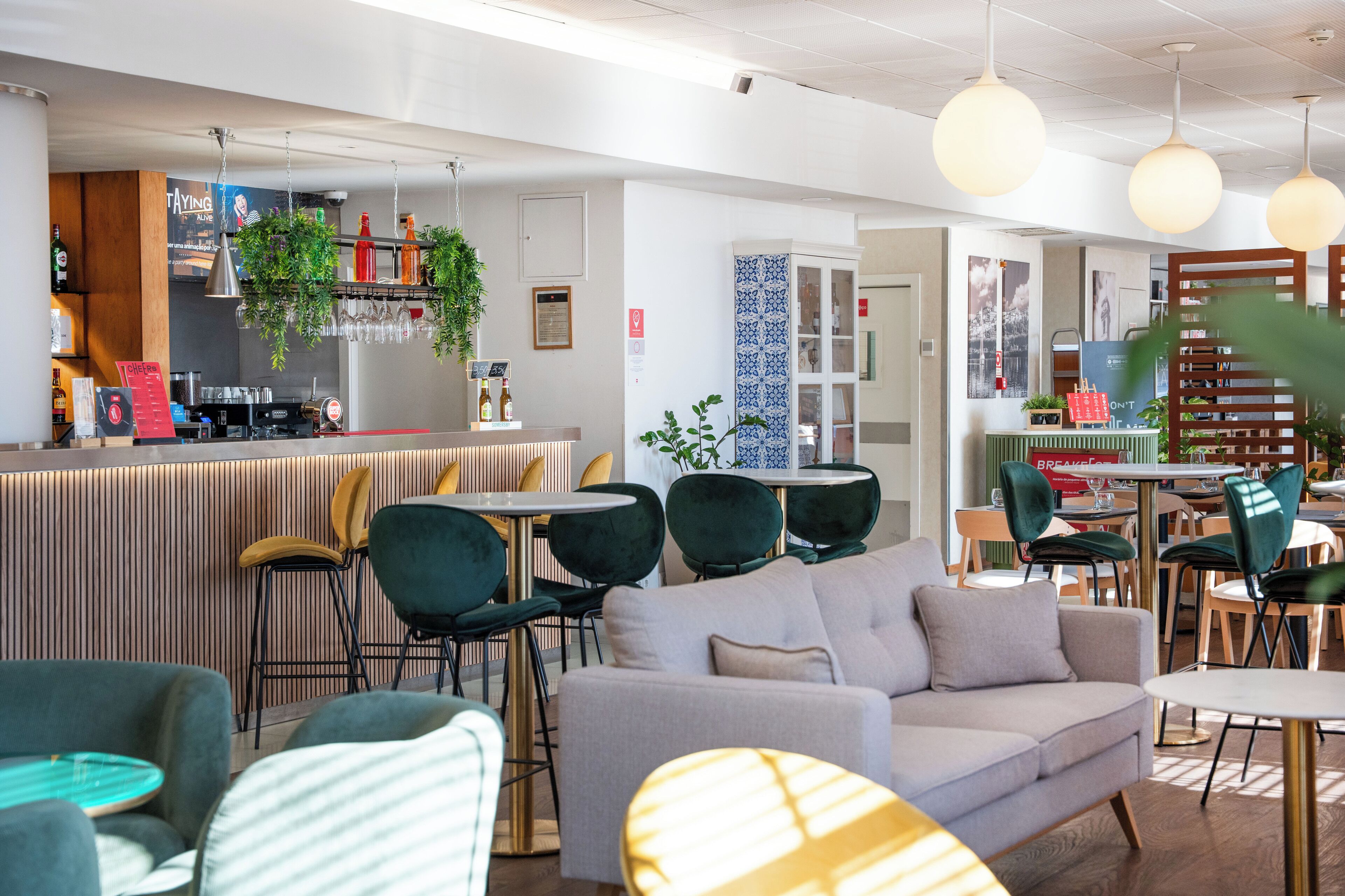Uma área de bar moderna com balcão, banquetas e plantas penduradas. Há áreas de estar com sofás e cadeiras, e uma escada com corrimão de madeira.