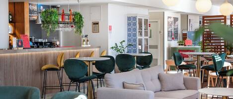 Uma área de bar moderna com balcão, banquetas e plantas penduradas. Há áreas de estar com sofás e cadeiras, e uma escada com corrimão de madeira.