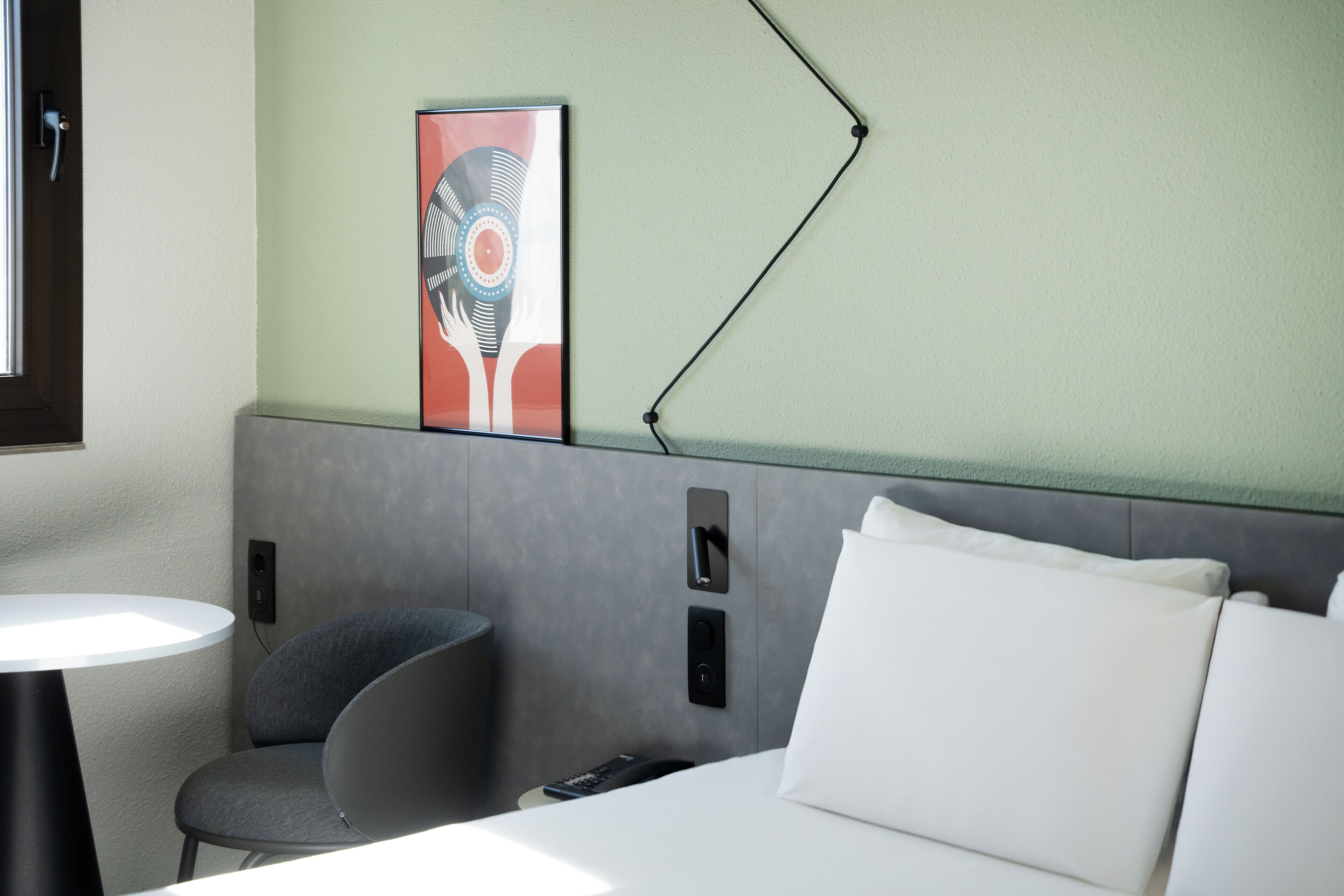 A hotel room with a bed, a bedside table, a chair, a wall-mounted fan, and a painting.