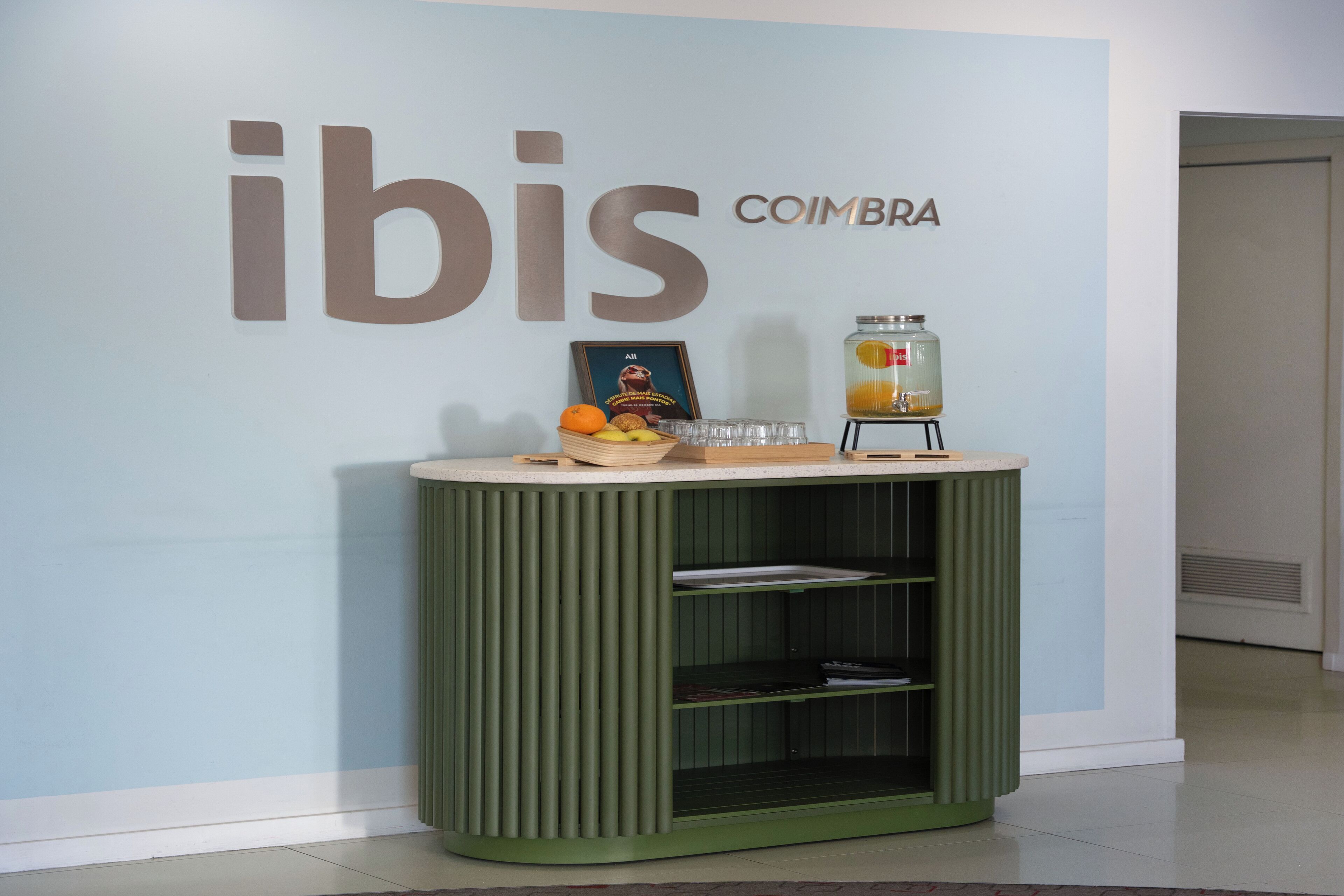 A green counter with a fruit bowl, a framed picture, and a drink dispenser against a wall with 'ibis' and 'COIMBRA' signage.