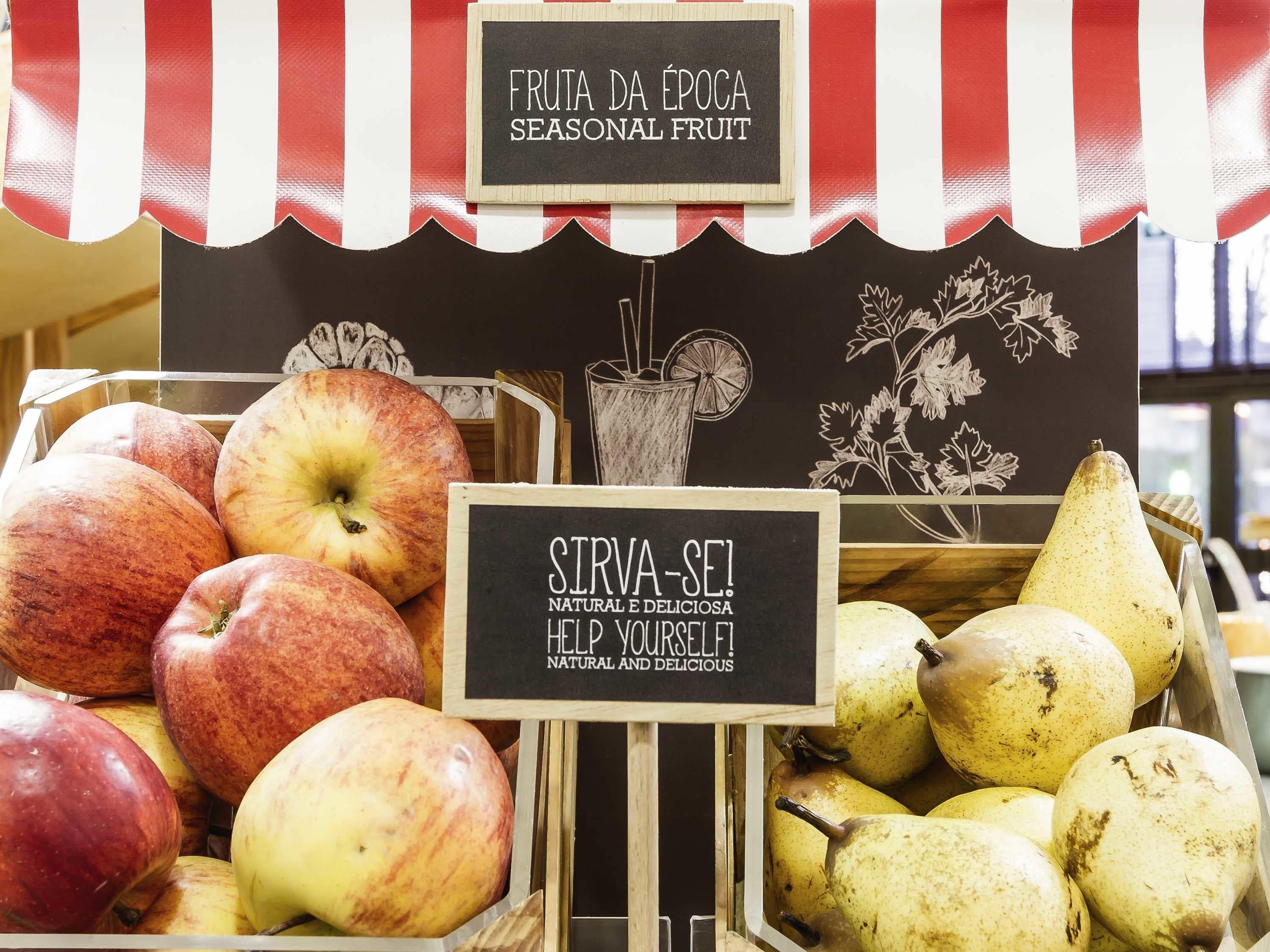 Un puesto de frutas con manzanas y peras, un cartel que dice 'FRUTA DA ÉPOCA SEASONAL FRUIT' y otro cartel que indica 'SIRVA-SE! NATURAL E DELICIOSA HELP YOURSELF! NATURAL AND DELICIOUS'.