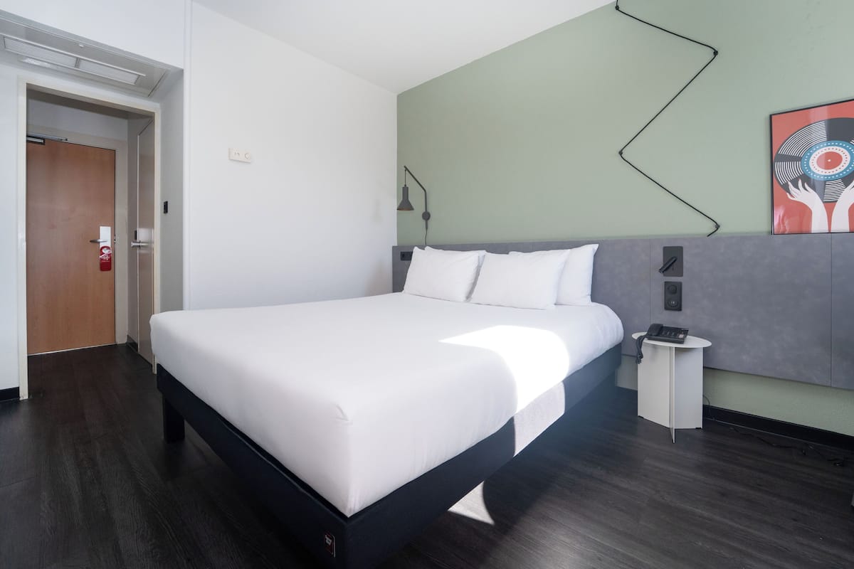 A modern hotel room with a neatly made bed, a bedside table with a phone, and a wall-mounted light fixture.
