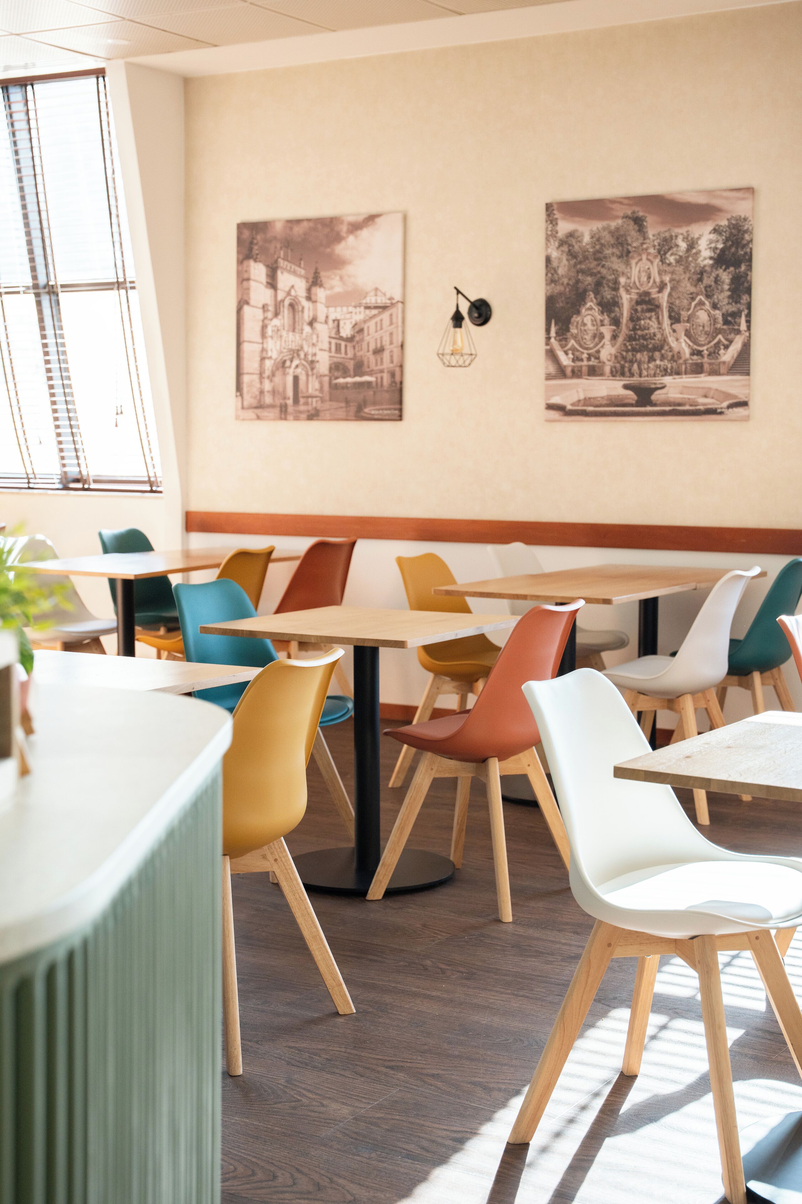 A dining area with wooden tables and colorful chairs, a wall-mounted light fixture, and two framed pictures.