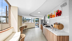 Une cuisine moderne avec un plan de travail blanc, des meubles en bois et une vue dégagée grâce à de grandes fenêtres. On aperçoit, au fond, des chaises en osier et un coin repas.