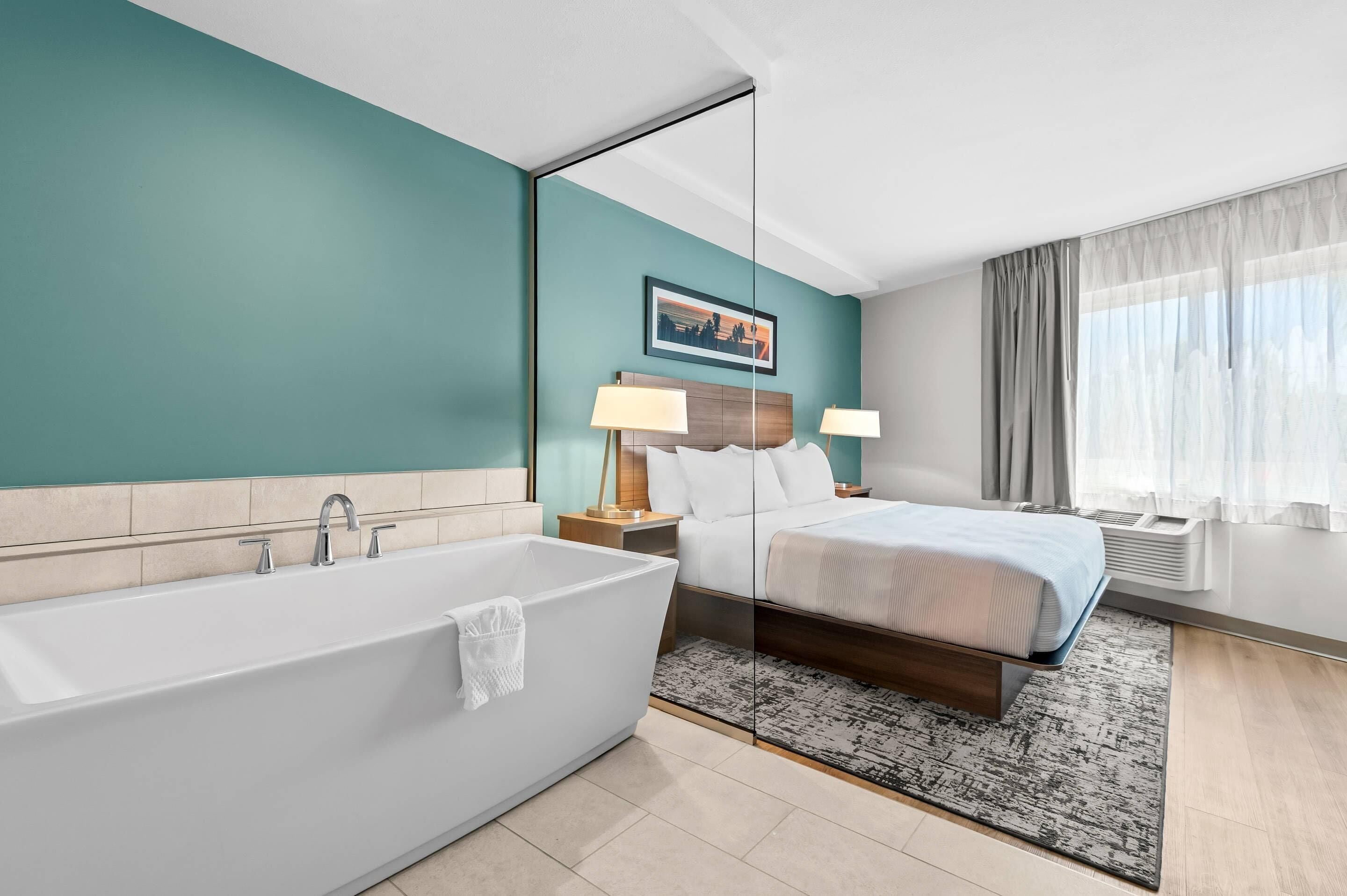 Una habitación de hotel moderna con bañera, cama, mesita de noche con lámparas, un cuadro enmarcado en la pared y una ventana con cortinas.