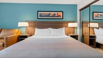 Une chambre d’hôtel avec un grand lit, deux tables de chevet avec des lampes, une tête de lit en bois, un fauteuil en cuir marron et un tableau encadré au mur.