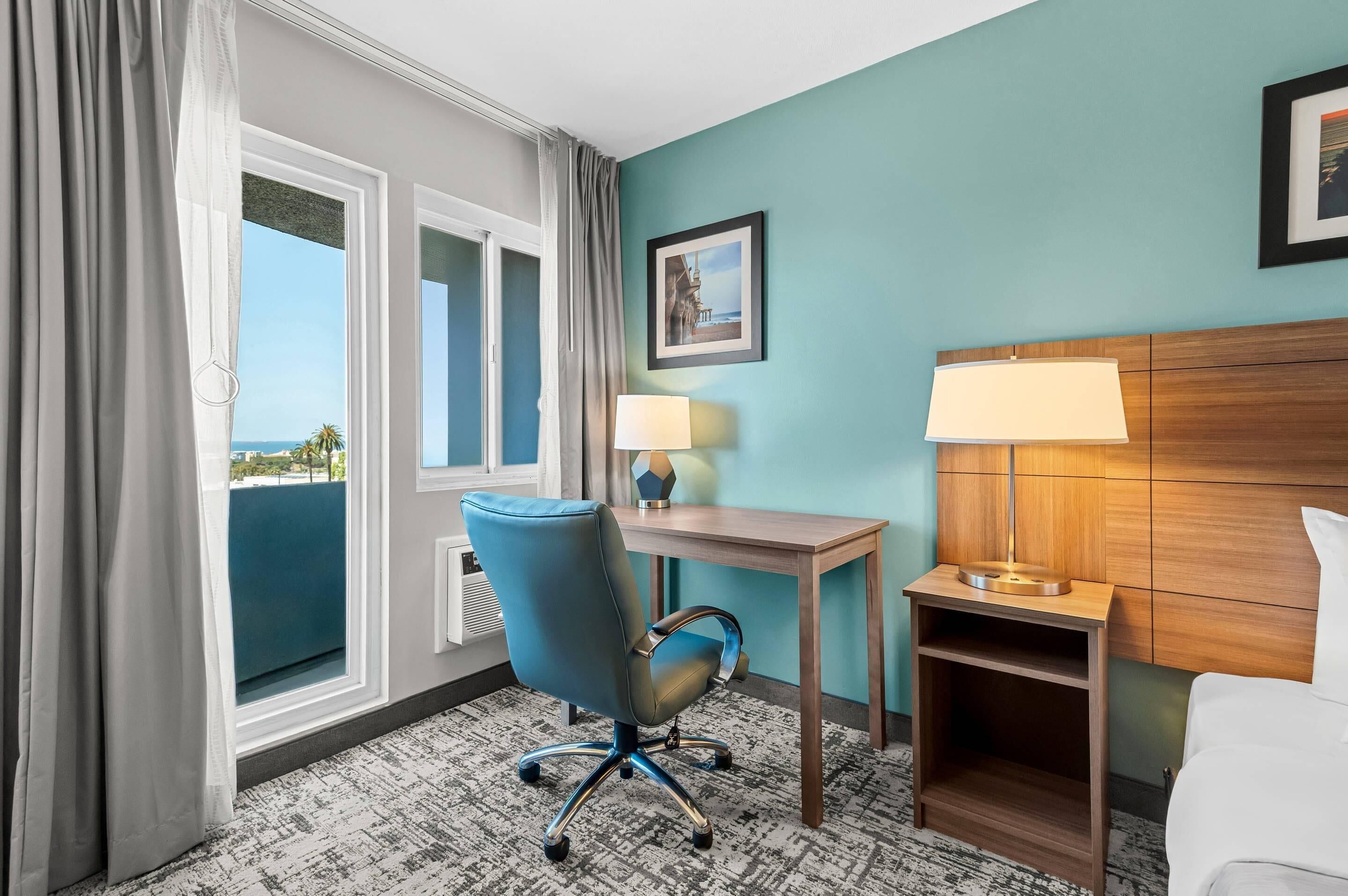 Een hotelkamer met een bureau, stoel, nachtkastje, lamp en een ingelijst schilderij aan de muur.
