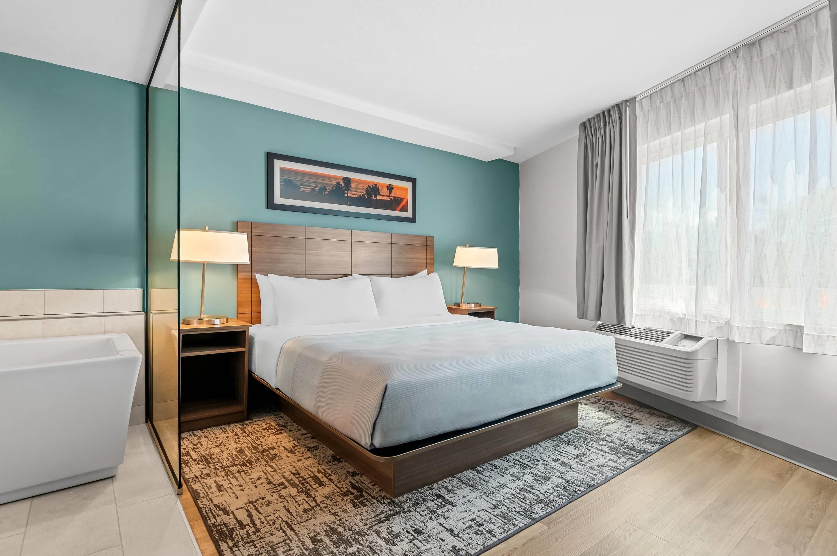 Hotelkamer met een bed, twee nachtkastjes met lampen, een groot raam met gordijnen, een wandairconditioner en een lijst met een afbeelding boven het bed.