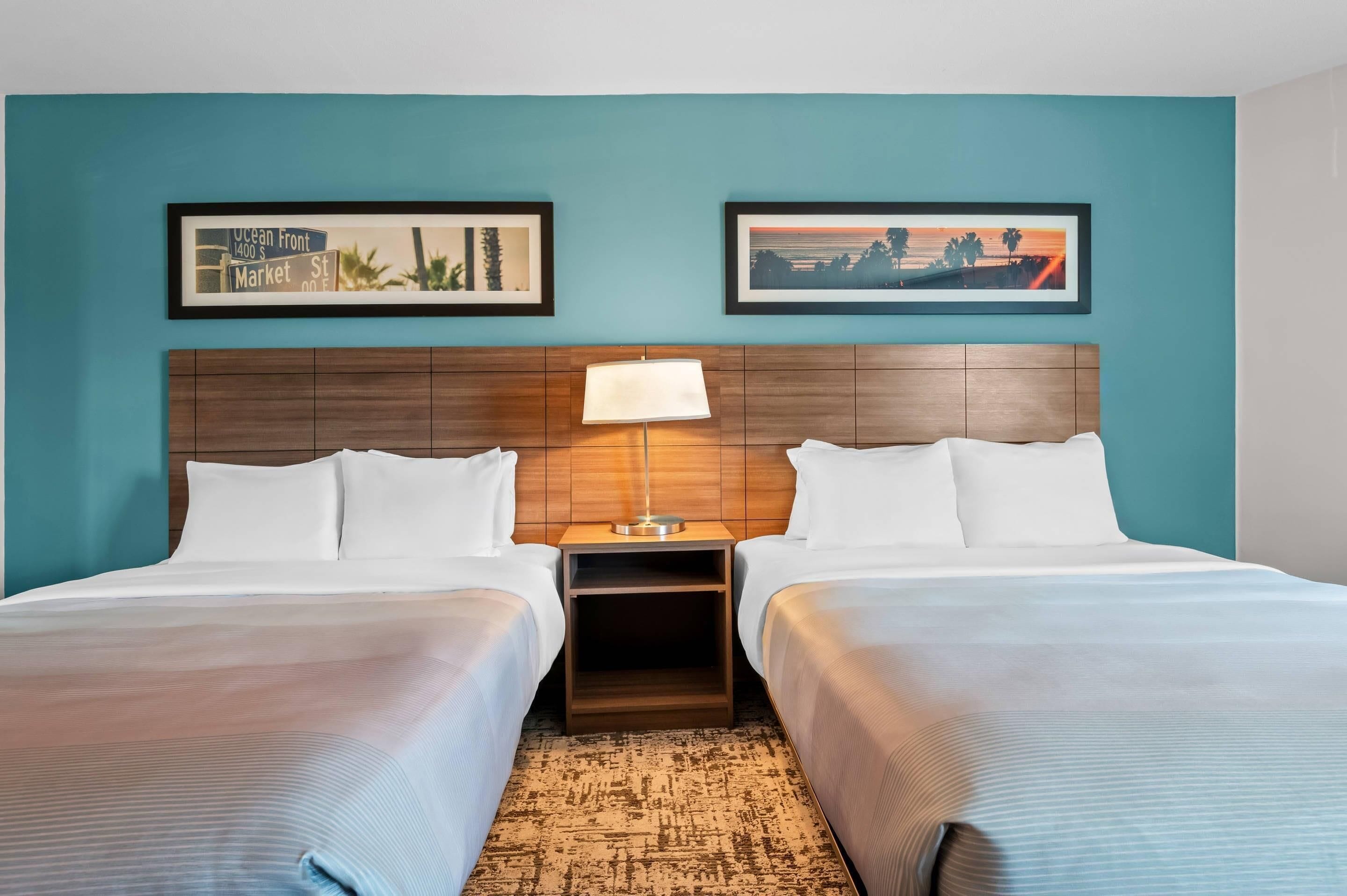 A hotel room with two beds, a wooden headboard, a nightstand with a lamp, and two framed pictures on the wall.