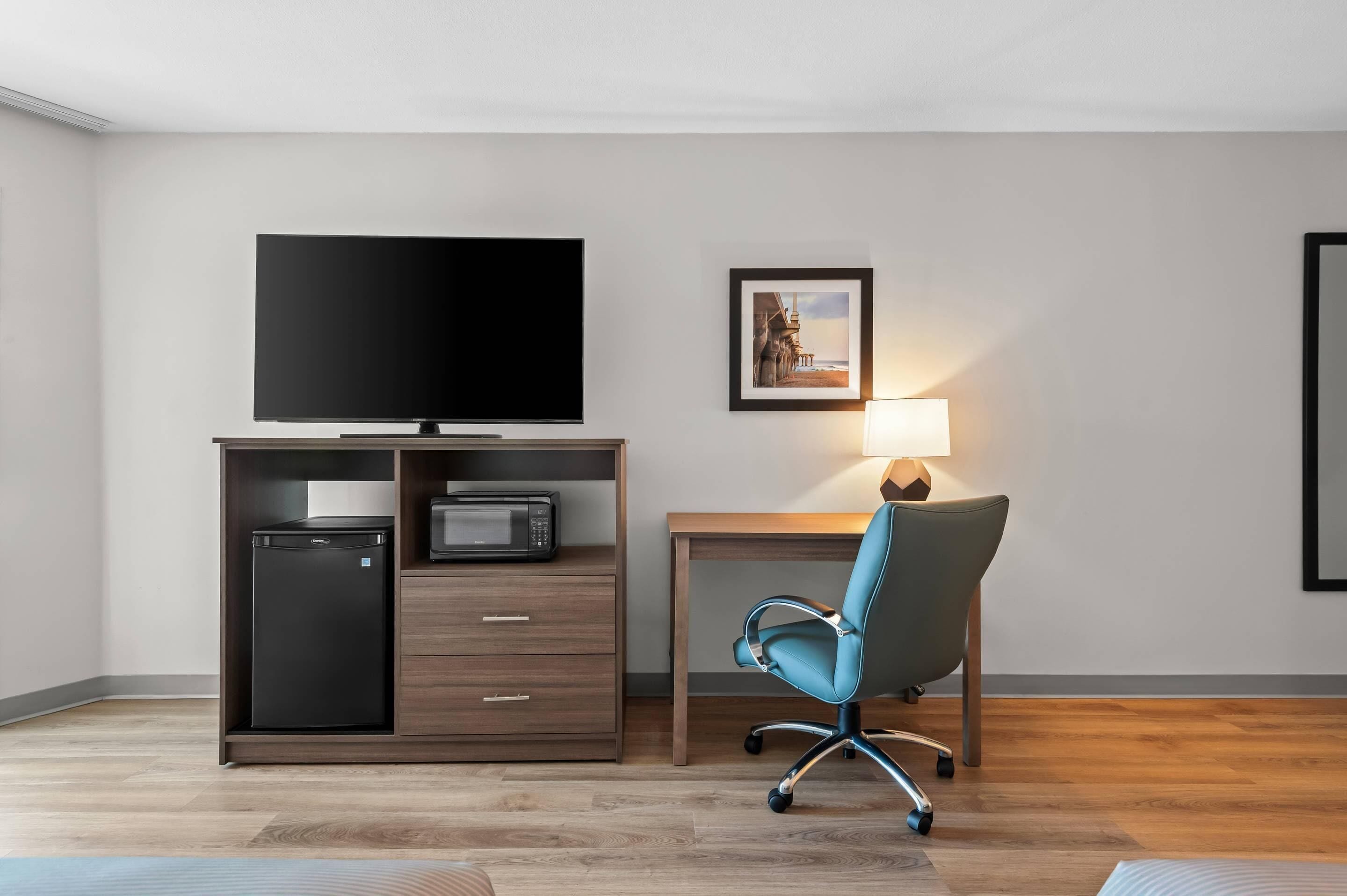 A hotel room with a flat-screen TV, a desk with a lamp, a chair, a small refrigerator, and a microwave.