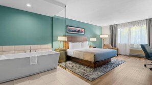 A modern hotel room with a large bathtub, a bed with a wooden headboard, two bedside lamps, a large window with curtains, and a painting on the wall. - Quality Inn & Suites Hermosa Beach (Hermosa Beach)