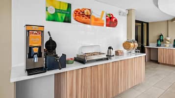 Una barra de desayuno con máquinas de gofres y café, una plancha para gofres y una presentación de fruta.