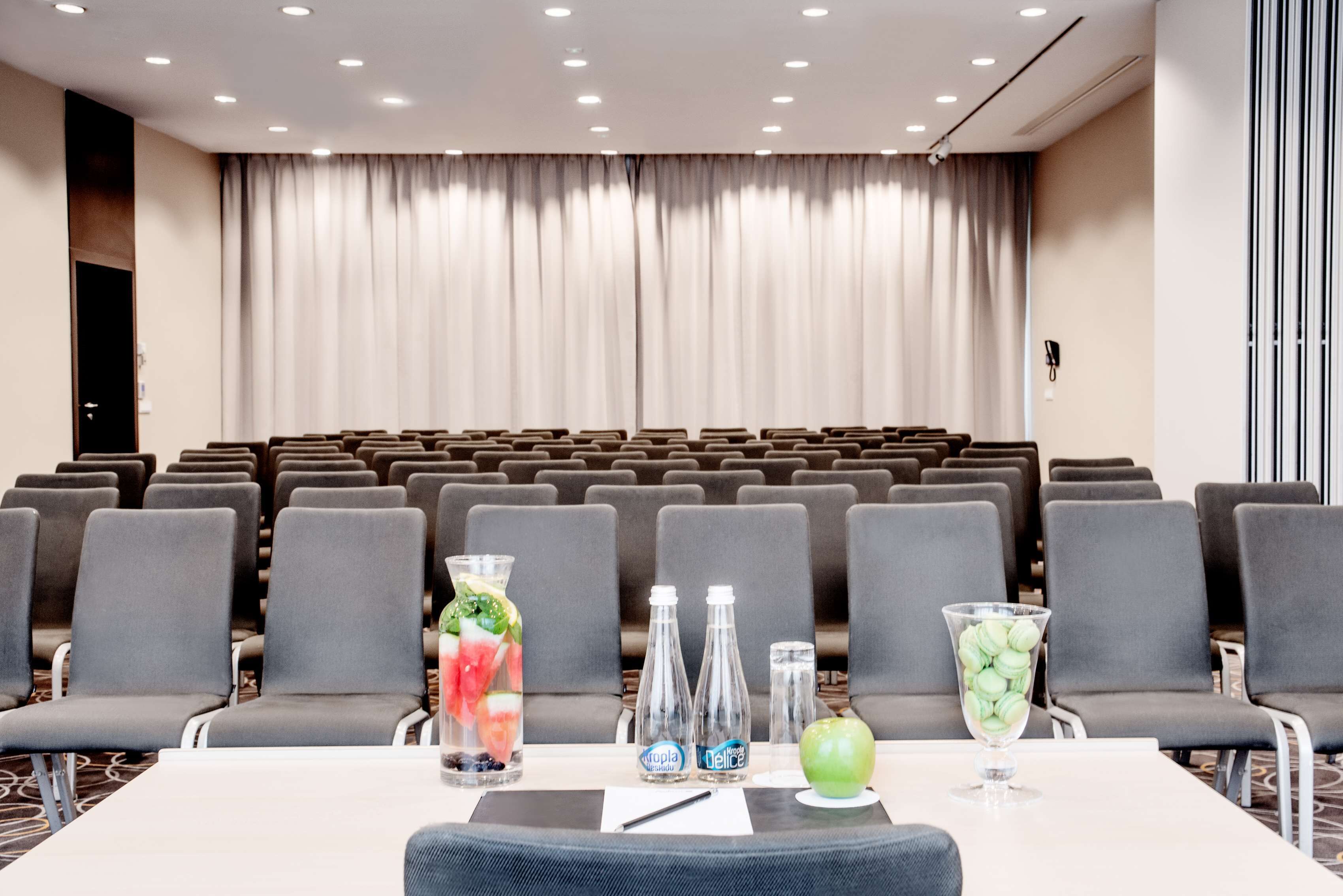 A conference room with rows of grey chairs, a table with water bottles, a glass of fruit, and a projector screen.