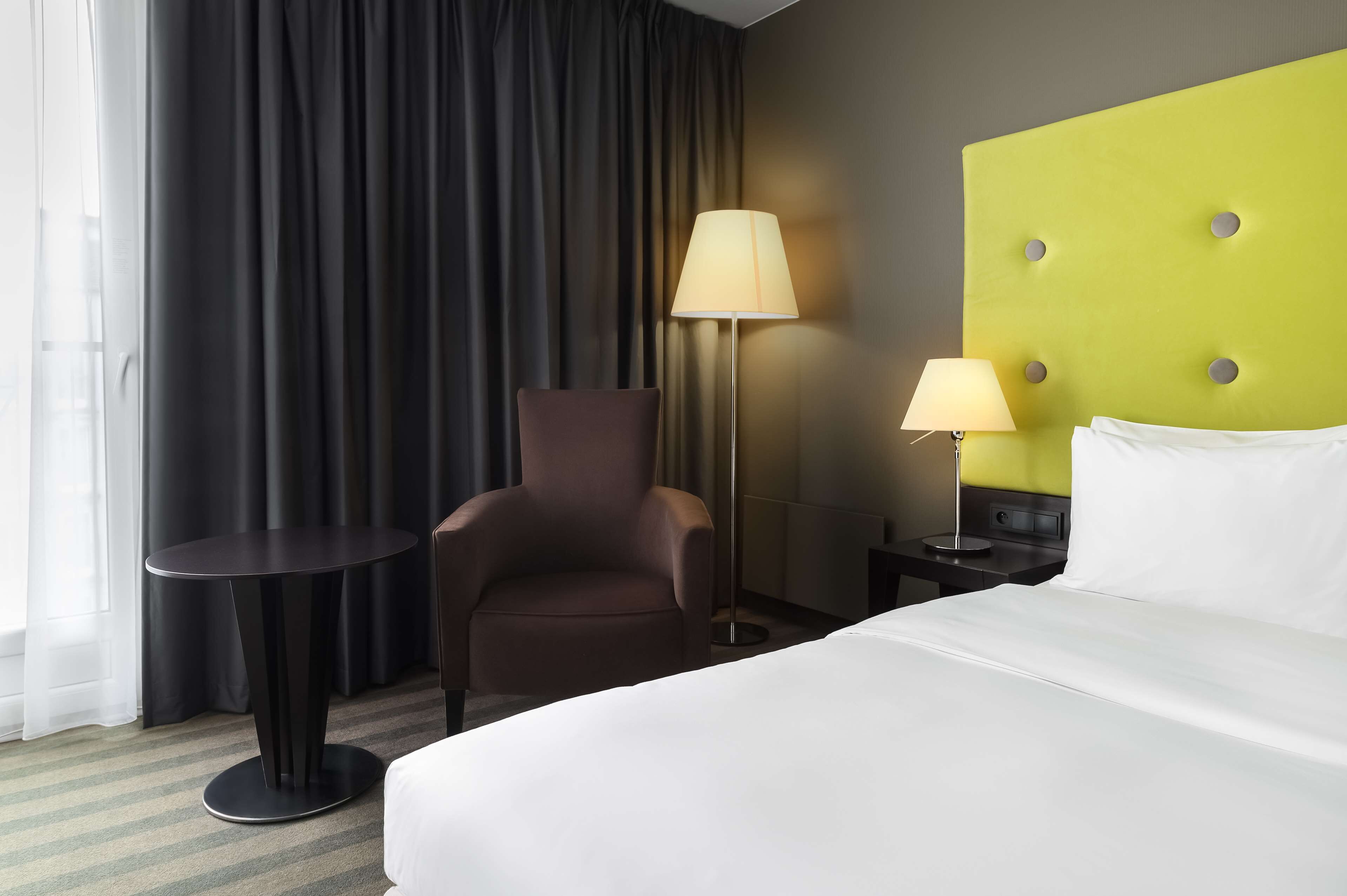 A hotel room with a bed, a chair, a table, and two lamps.