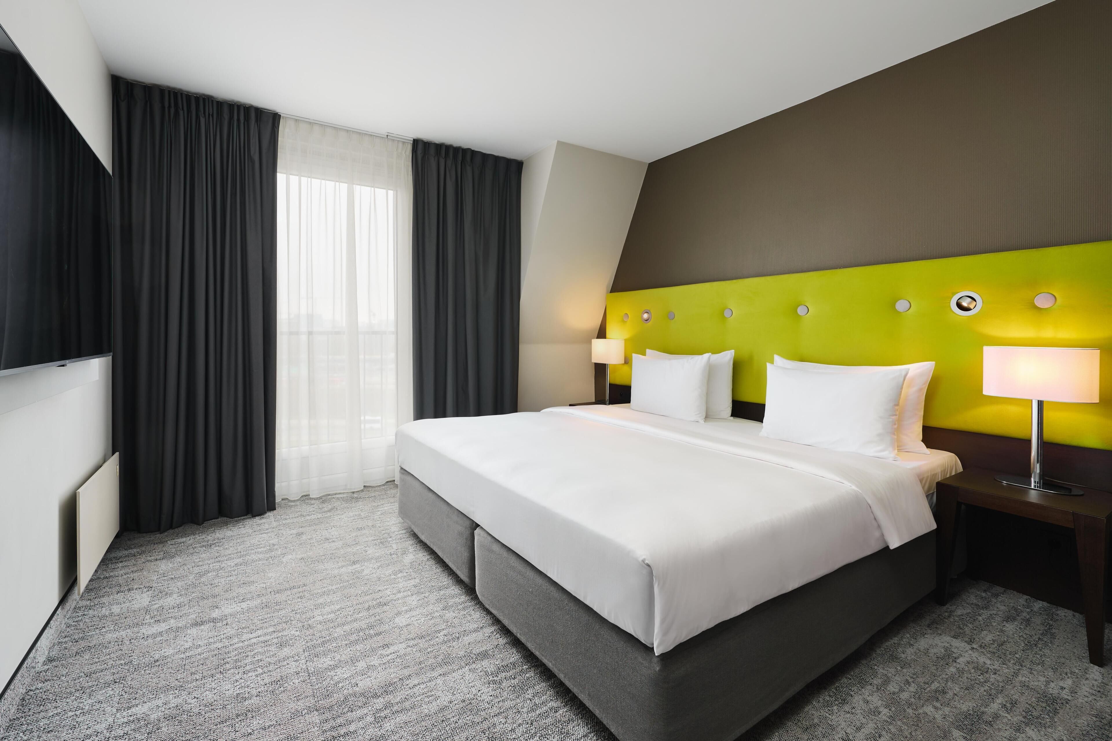 A hotel room with a large bed, a flat-screen TV, a bedside table with a lamp, and a window with curtains.