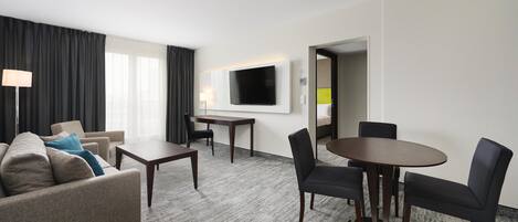 A hotel room with a sofa, a dining table with chairs, a flat-screen TV mounted on the wall, and a carpeted floor.