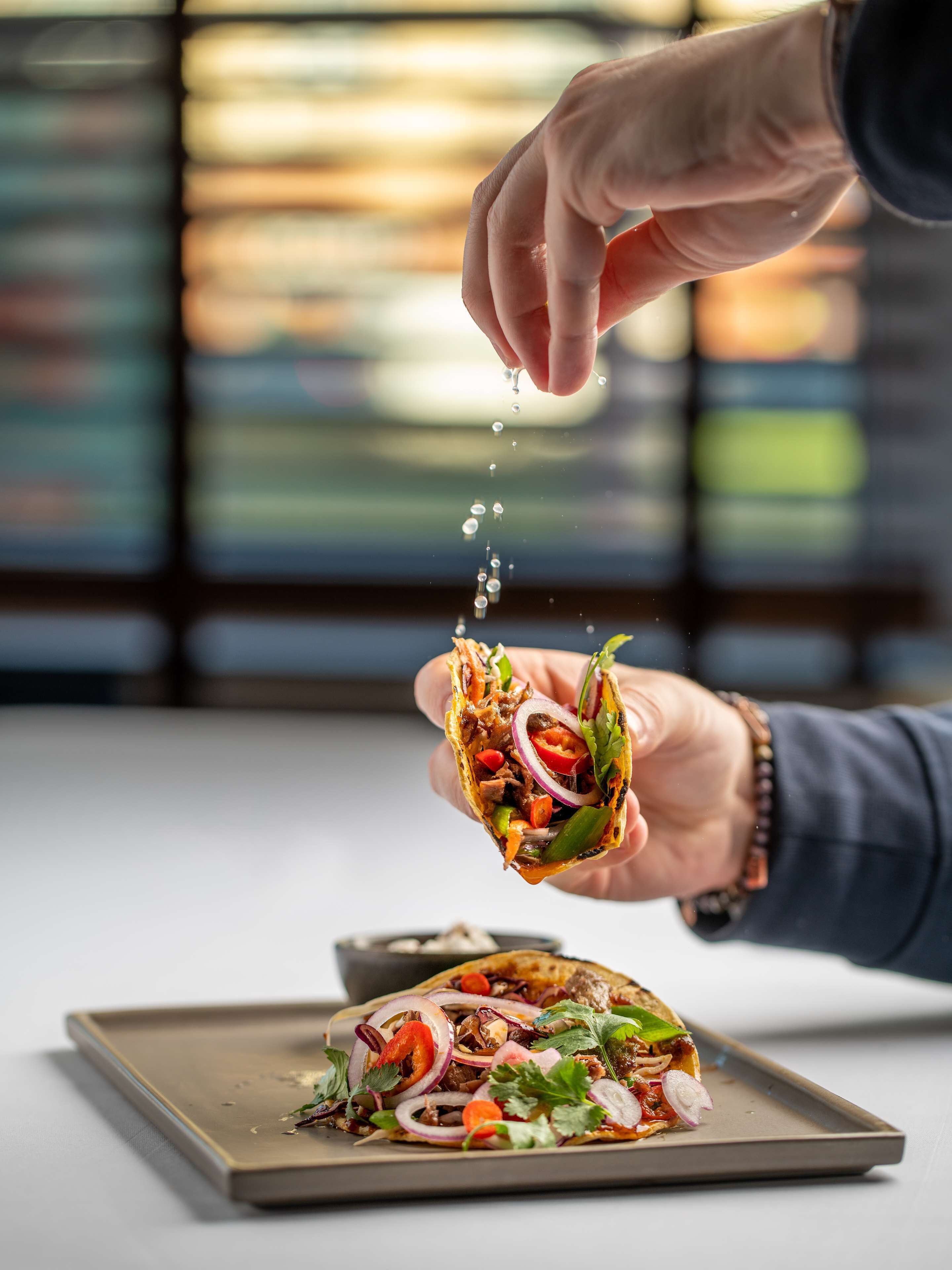 A hand holding a taco with various toppings, with a plate of similar tacos in the background.