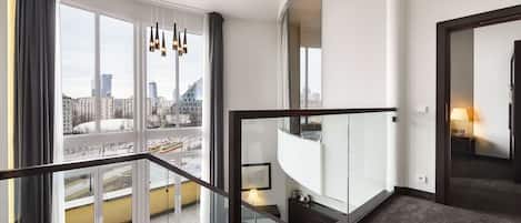Ett modernt hotellrum med stora fönster som erbjuder utsikt över staden, ett glasräcke på balkongen och en trappa som leder upp till en våning ovanför.