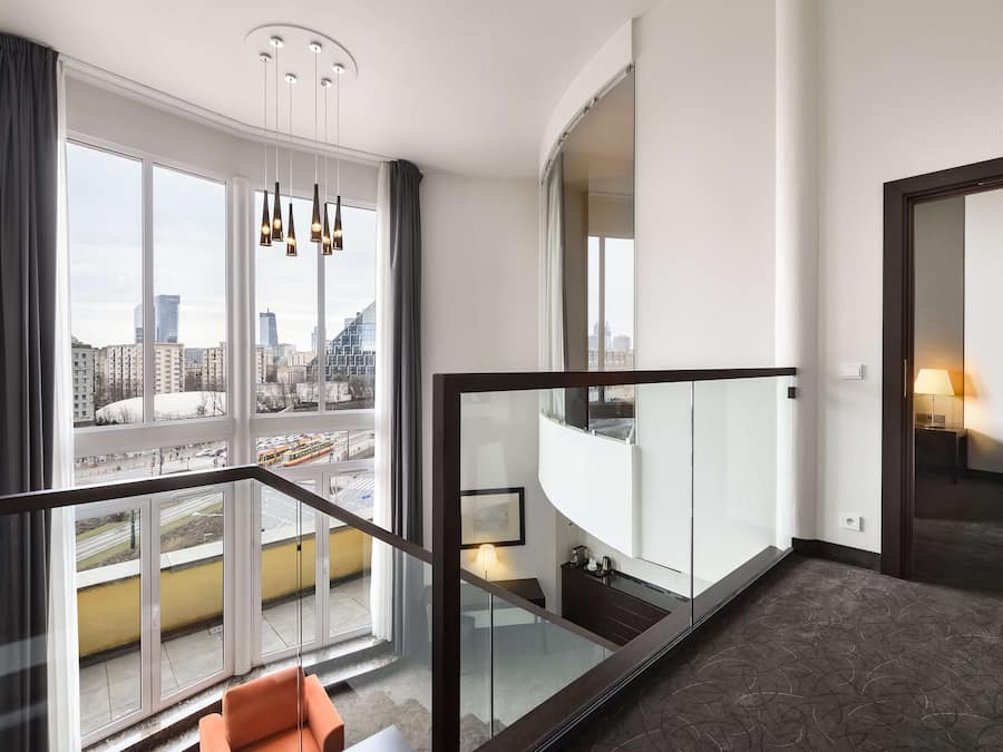 A modern hotel room with large windows offering city views, a glass railing on the balcony, and a staircase leading to an upper floor.