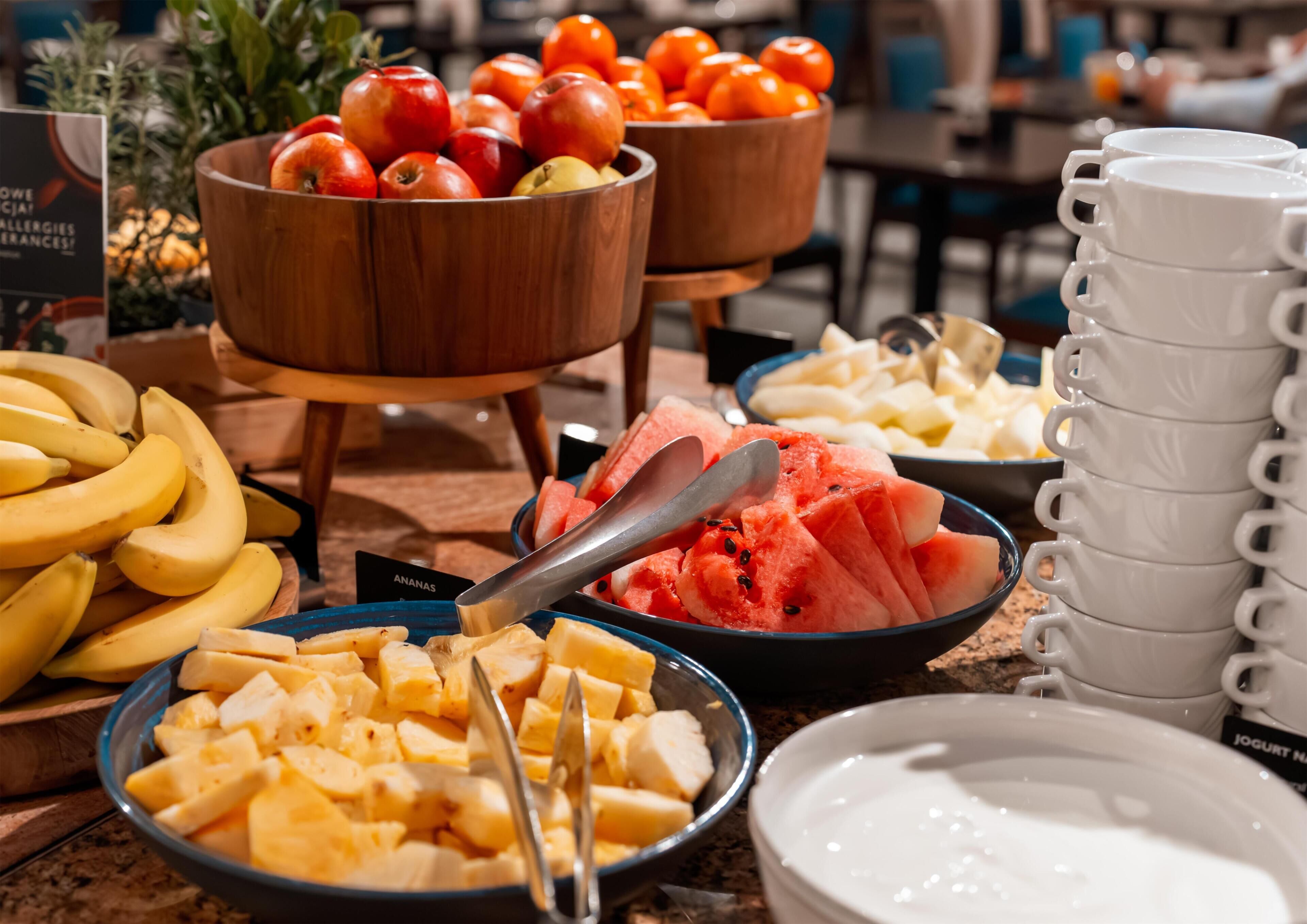 Un bufé con diversas frutas y una pila de tazas blancas.