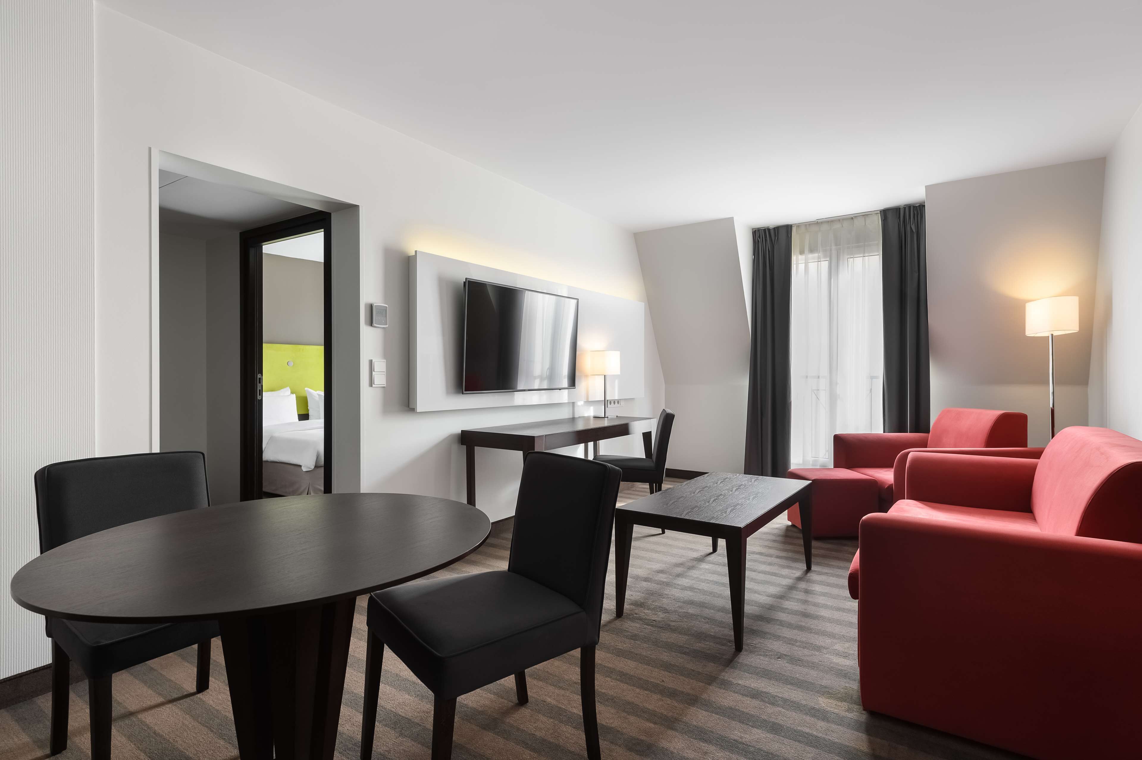 Una habitación de hotel con una mesa de comedor, sillas, un sofá rojo, un televisor montado en la pared y una lámpara.