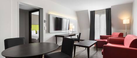 Una habitación de hotel con una mesa de comedor, sillas, un sofá rojo, un televisor montado en la pared y una lámpara.