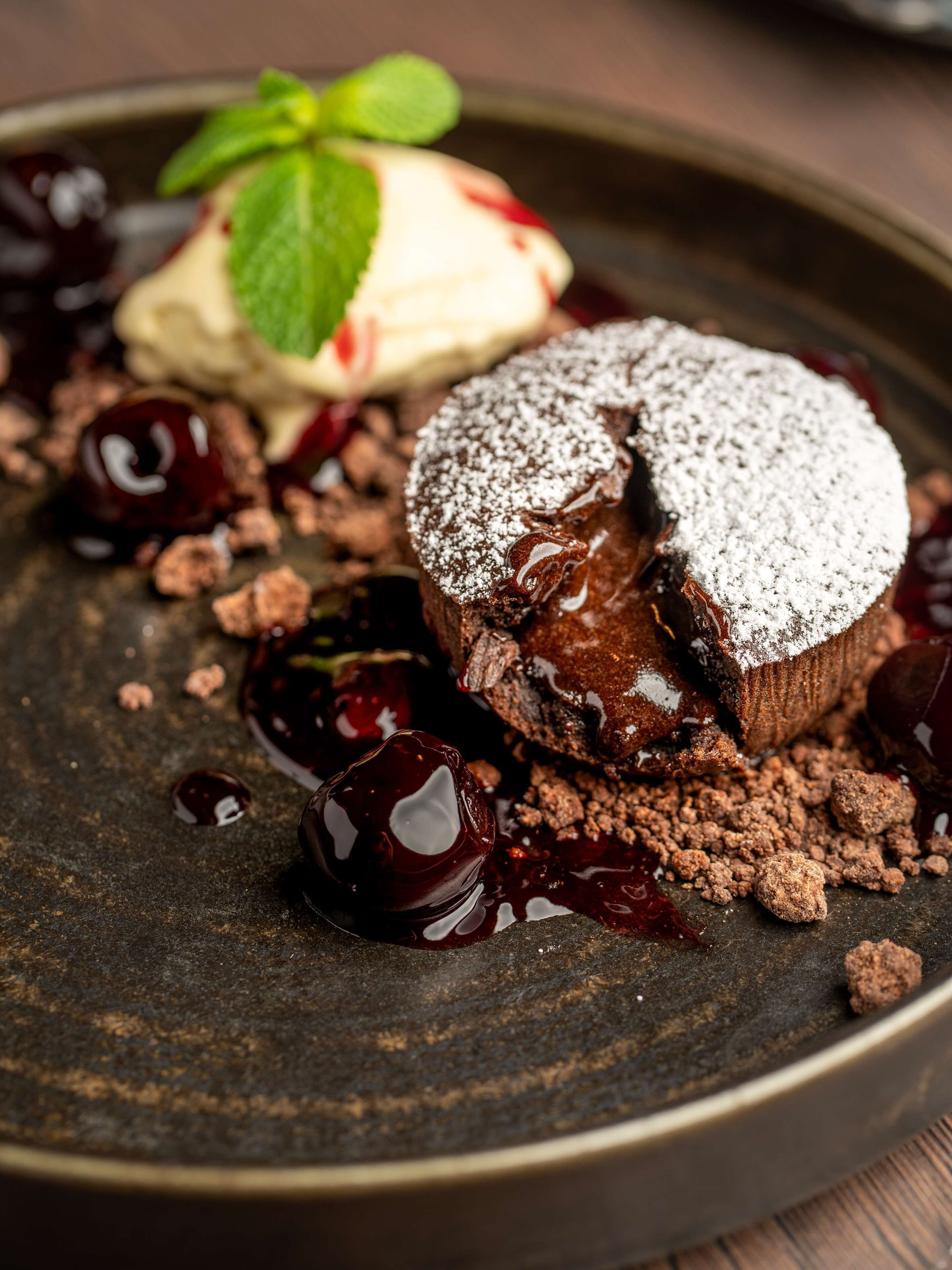 Un postre con pastel de chocolate, cerezas y una hoja de menta sobre un plato oscuro.