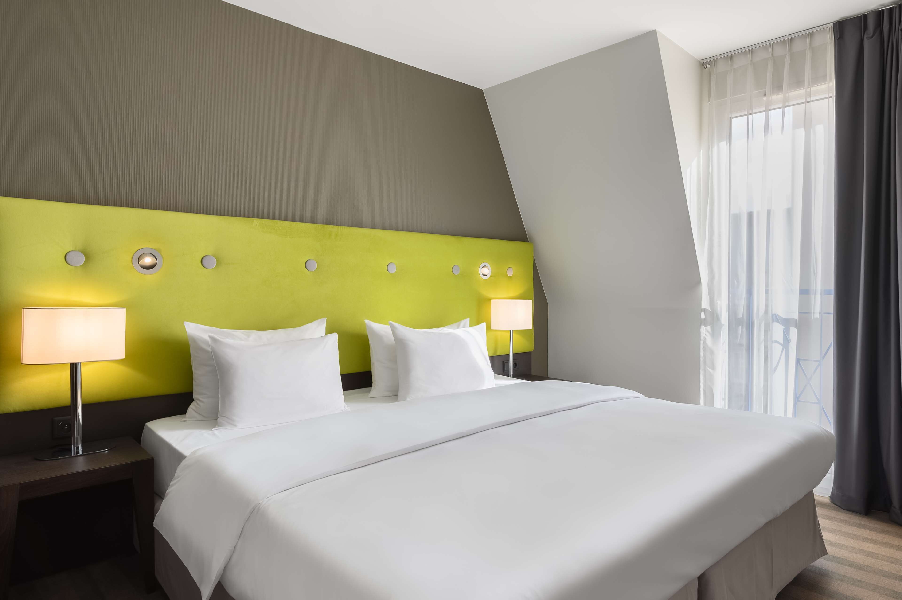 A hotel room with a large bed, two bedside tables with lamps, a green accent wall, and a view of the outdoors.