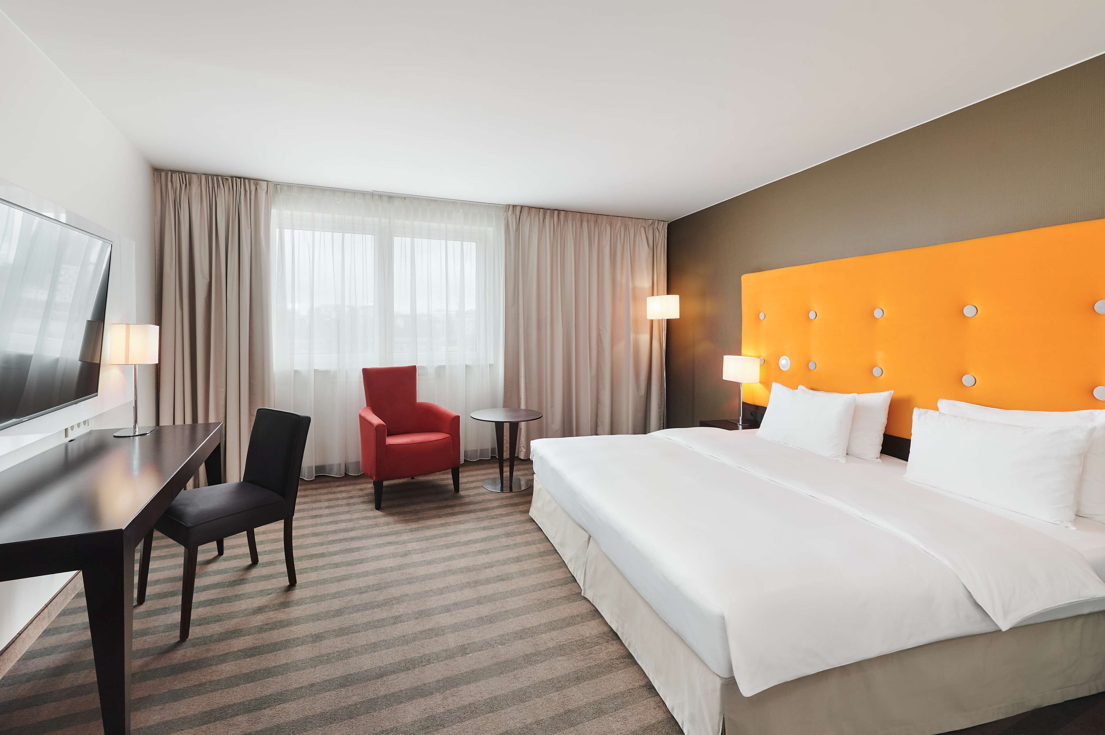 Une chambre d’hôtel avec un grand lit, un bureau avec une chaise, un fauteuil rouge, une petite table et un téléviseur.
