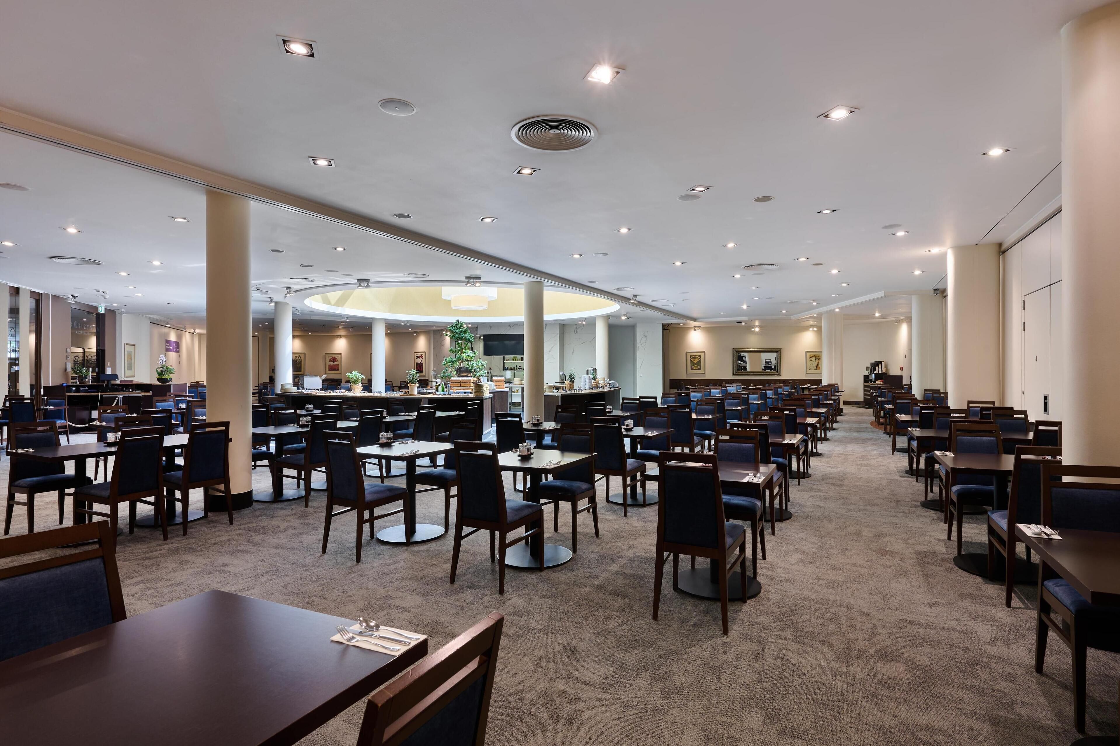 A spacious hotel dining area with multiple tables and chairs, a central column, and a circular ceiling design.