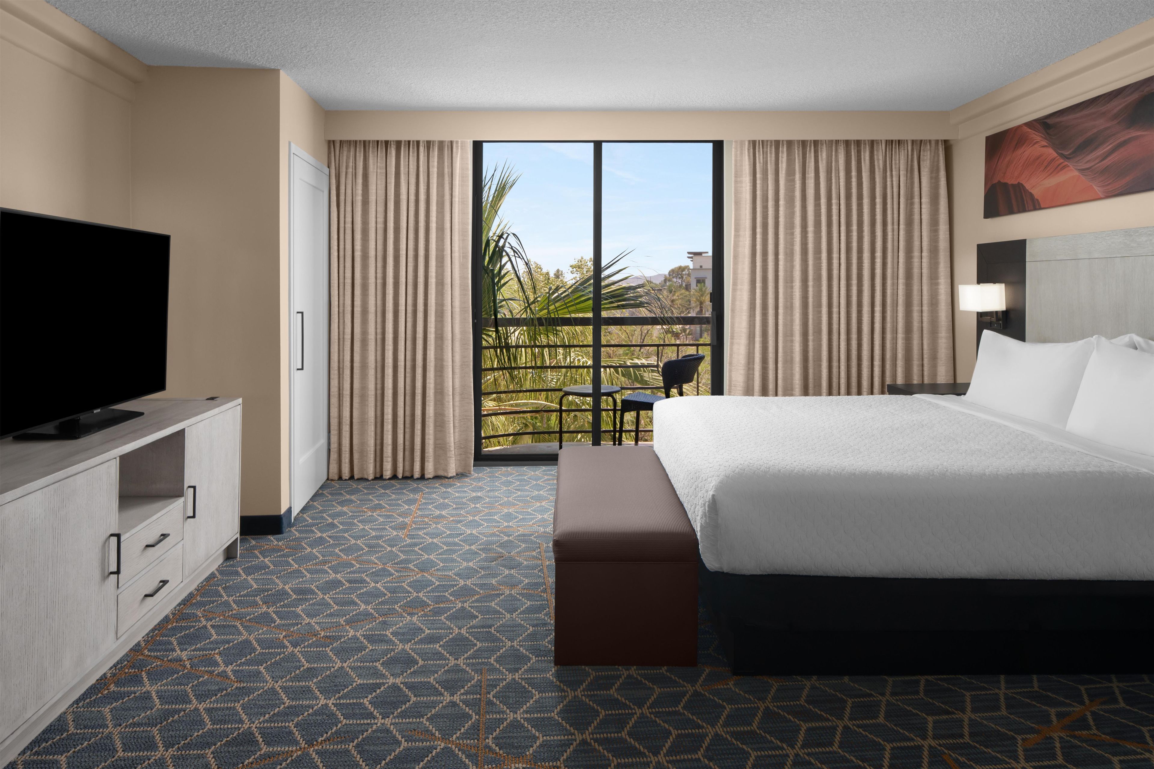 A hotel room with a large bed, a flat-screen TV, a desk with drawers, a window with curtains, and a view of greenery outside.
