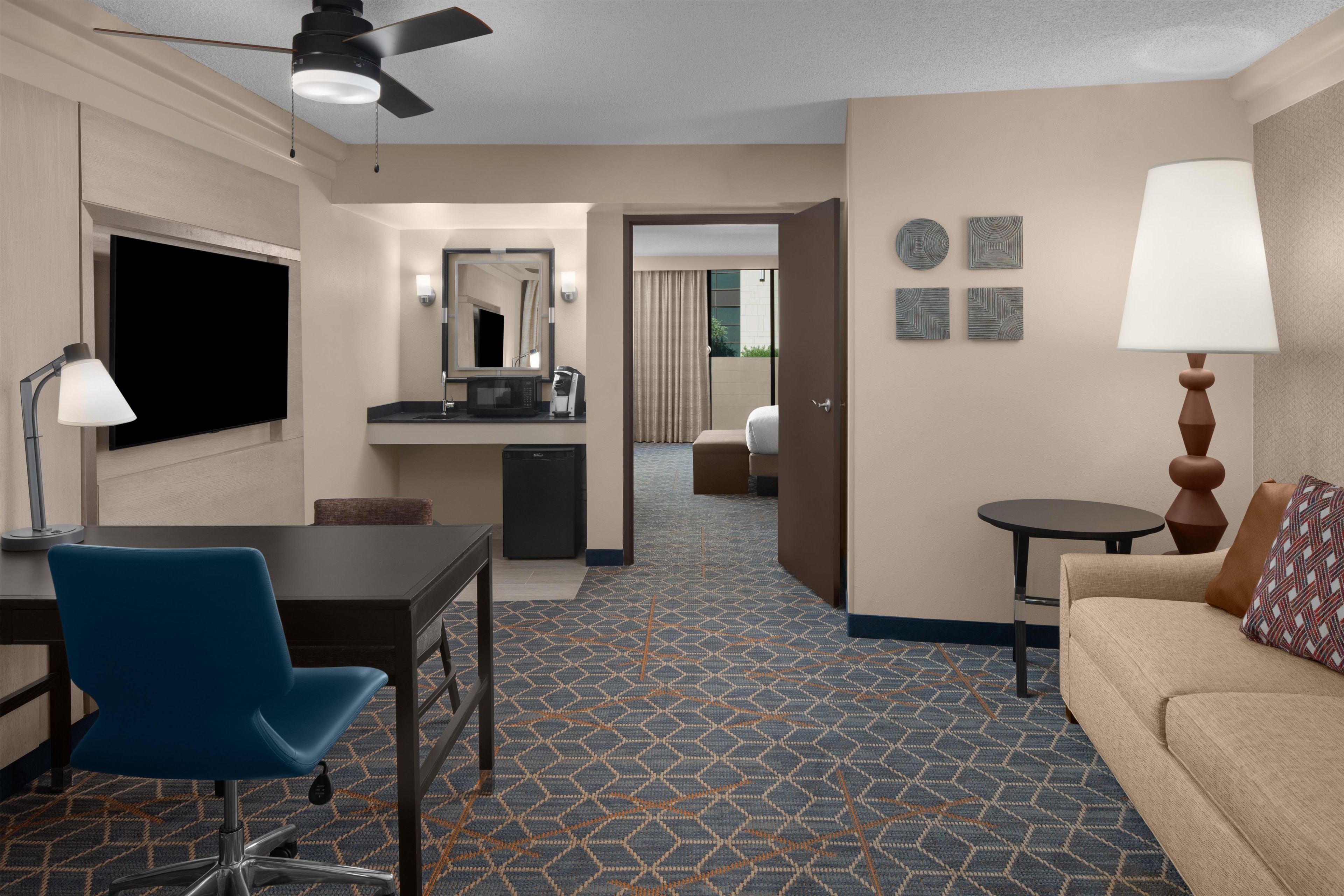 Habitación de hotel con televisor de pantalla plana, escritorio con silla, sofá, mesita con lámpara, microondas y refrigerador pequeño.