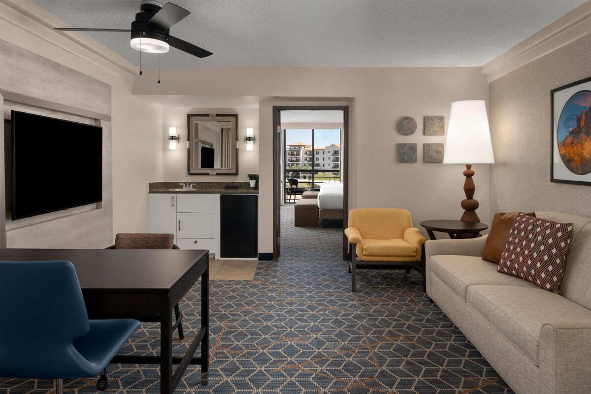 A hotel room with a flat-screen TV, a ceiling fan, a sofa, a chair, a small table, and a patterned floor.