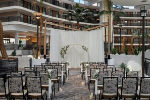 A multi-level atrium with a central aisle leading to a white archway, surrounded by rows of chairs and palm trees.