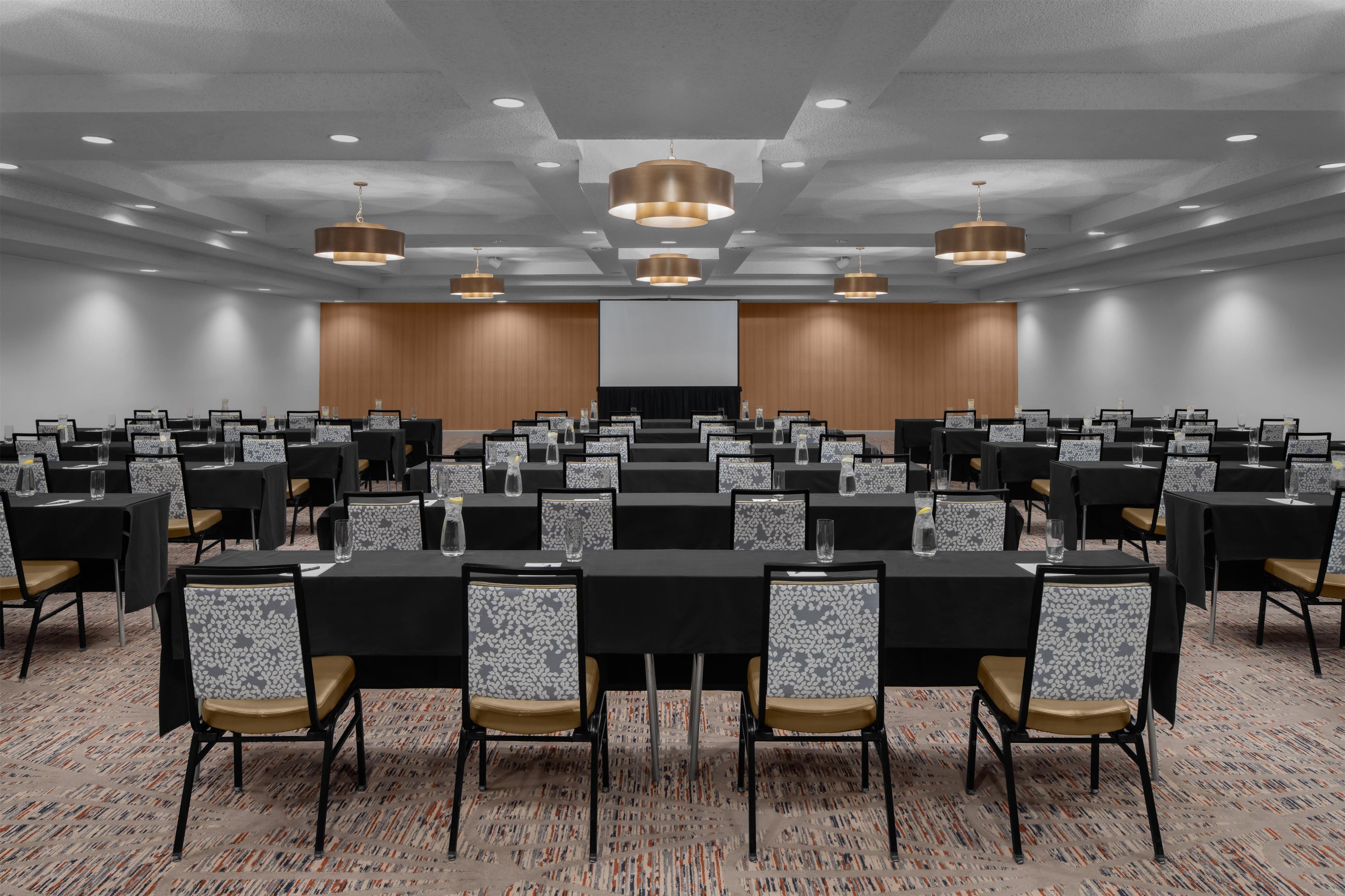 A conference room with multiple tables and chairs, equipped with overhead lighting and a projection screen.