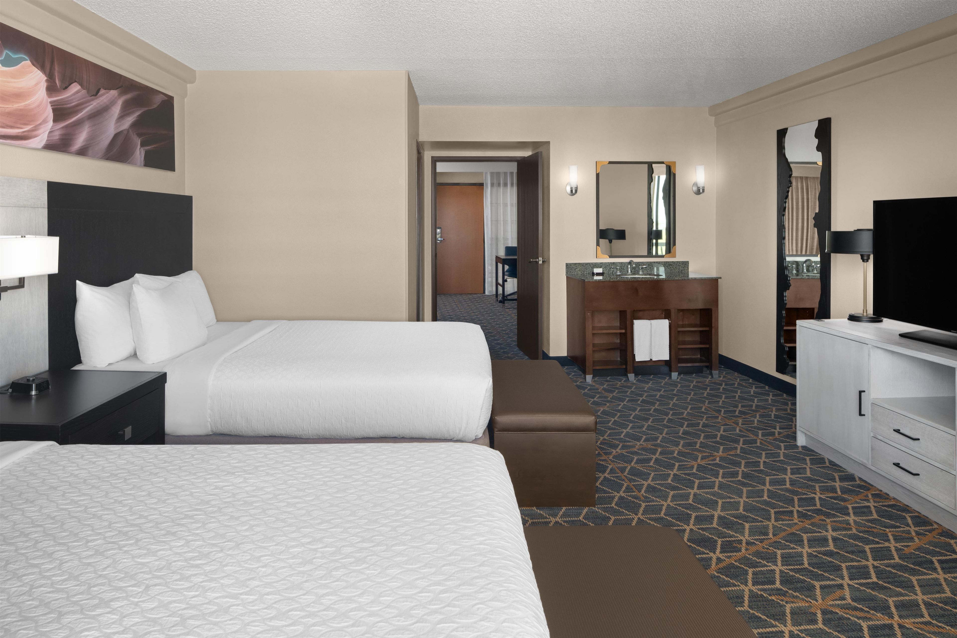 A hotel room with two beds, a desk, a dresser, a TV, and a bathroom with a mirror and sink.