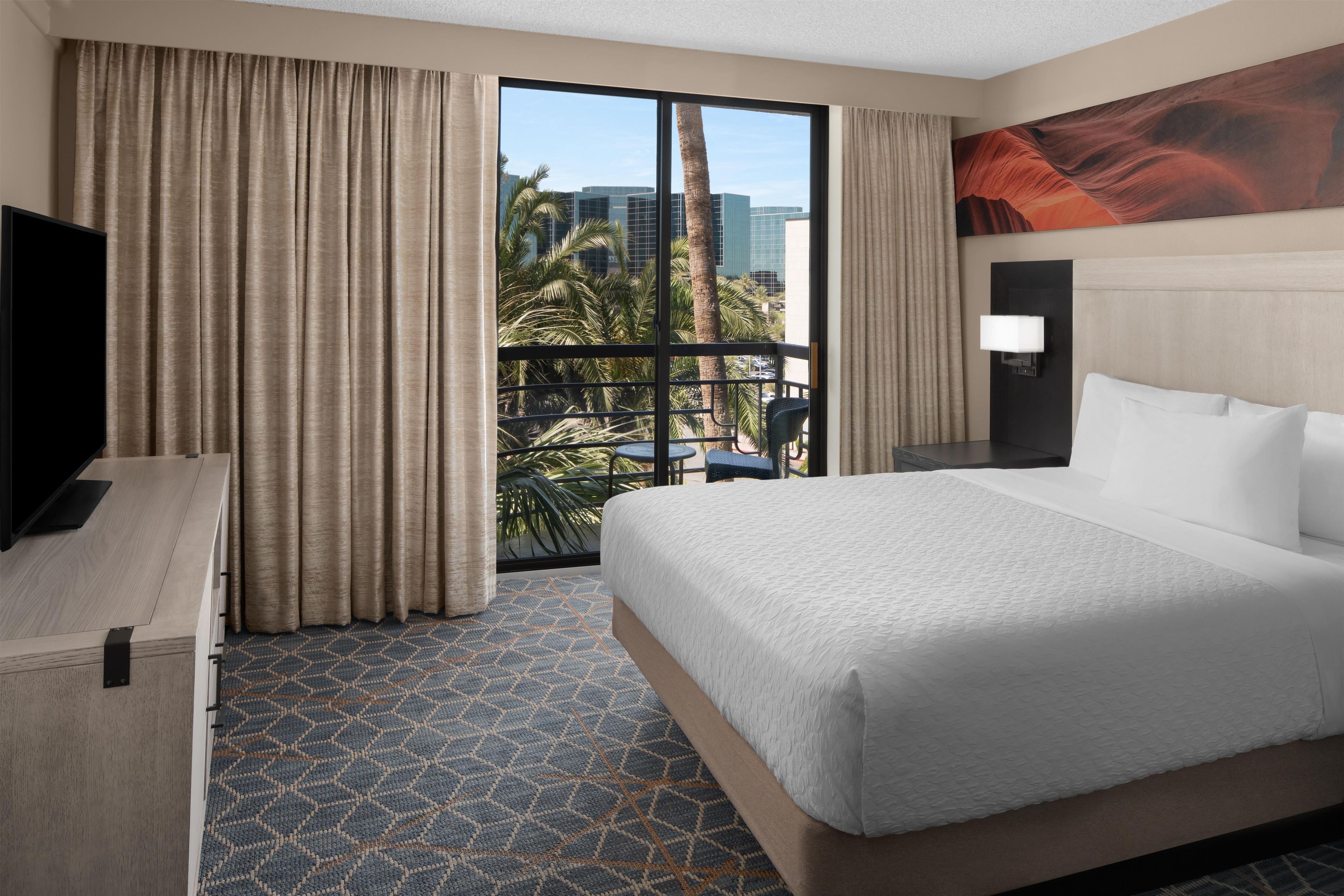 Habitación de hotel con una cama grande, un escritorio, un televisor y vistas a palmeras y edificios a través de la ventana.