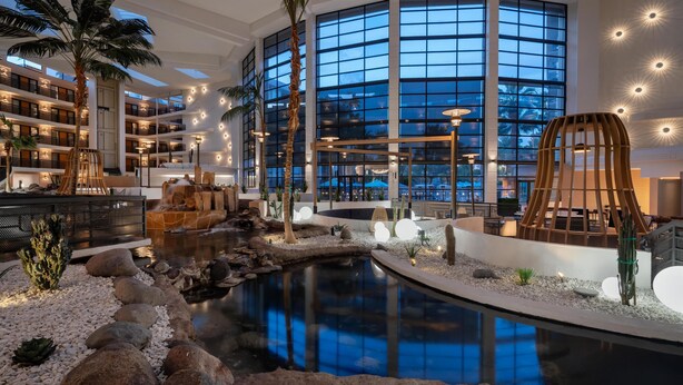 Un interior de hotel moderno con una amplia piscina cubierta, palmeras y vistas de la ciudad a través de grandes ventanales.