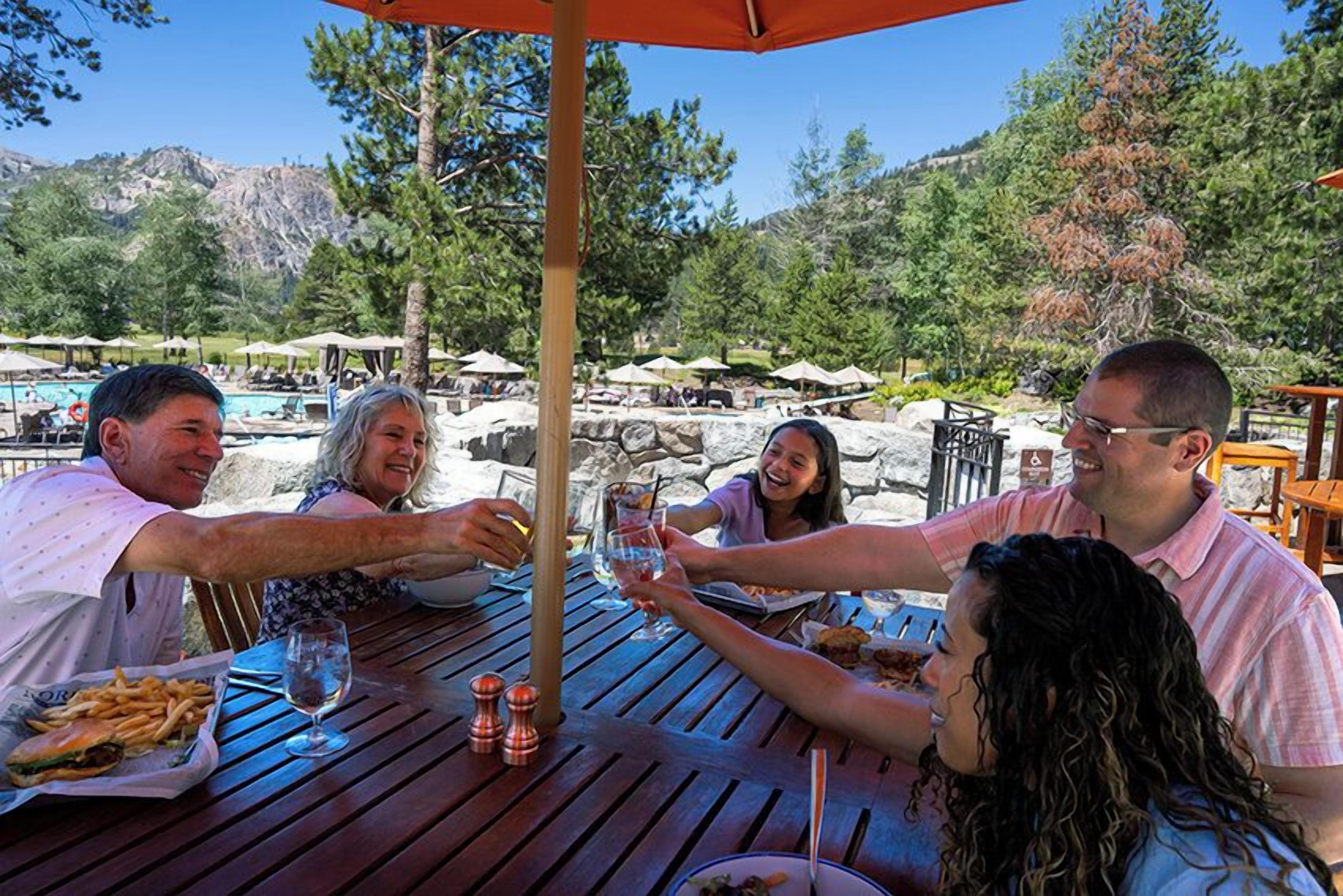 Un grupo de personas brindando en una mesa al aire libre, con vista a una piscina y montañas al fondo.