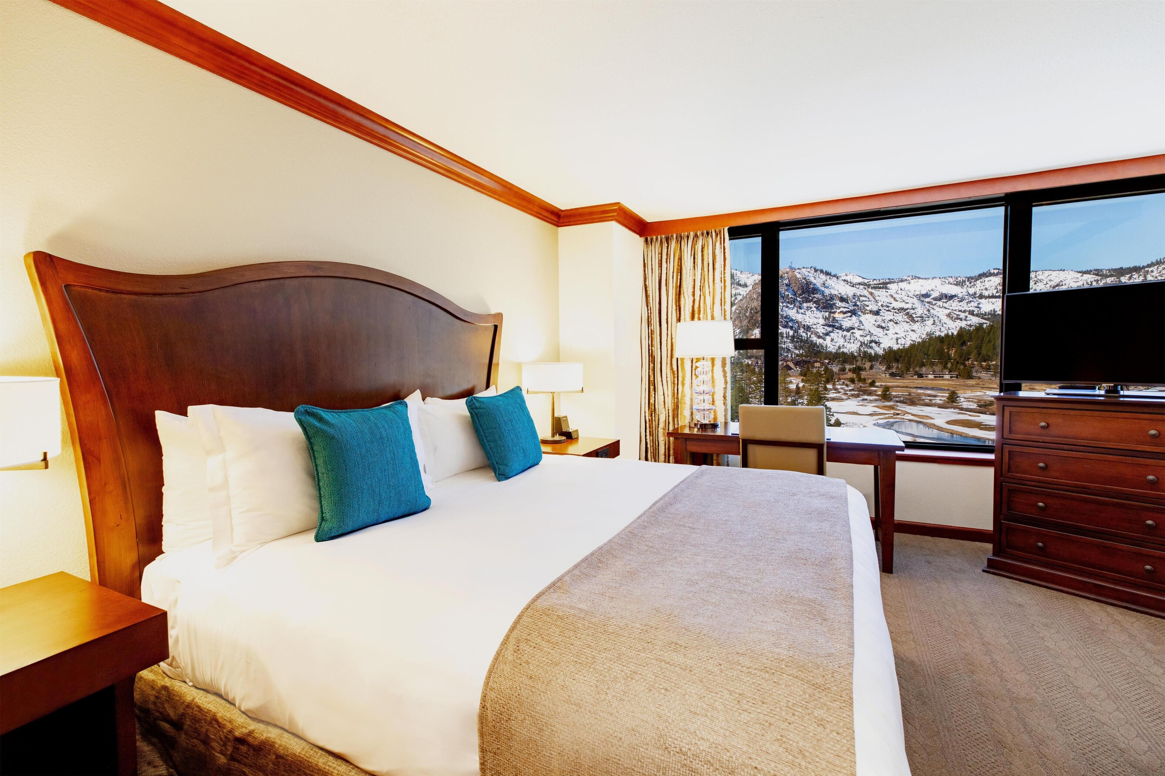 Ett hotellrum med en stor säng, ett skrivbord, en byrå och utsikt över snöklädda berg.