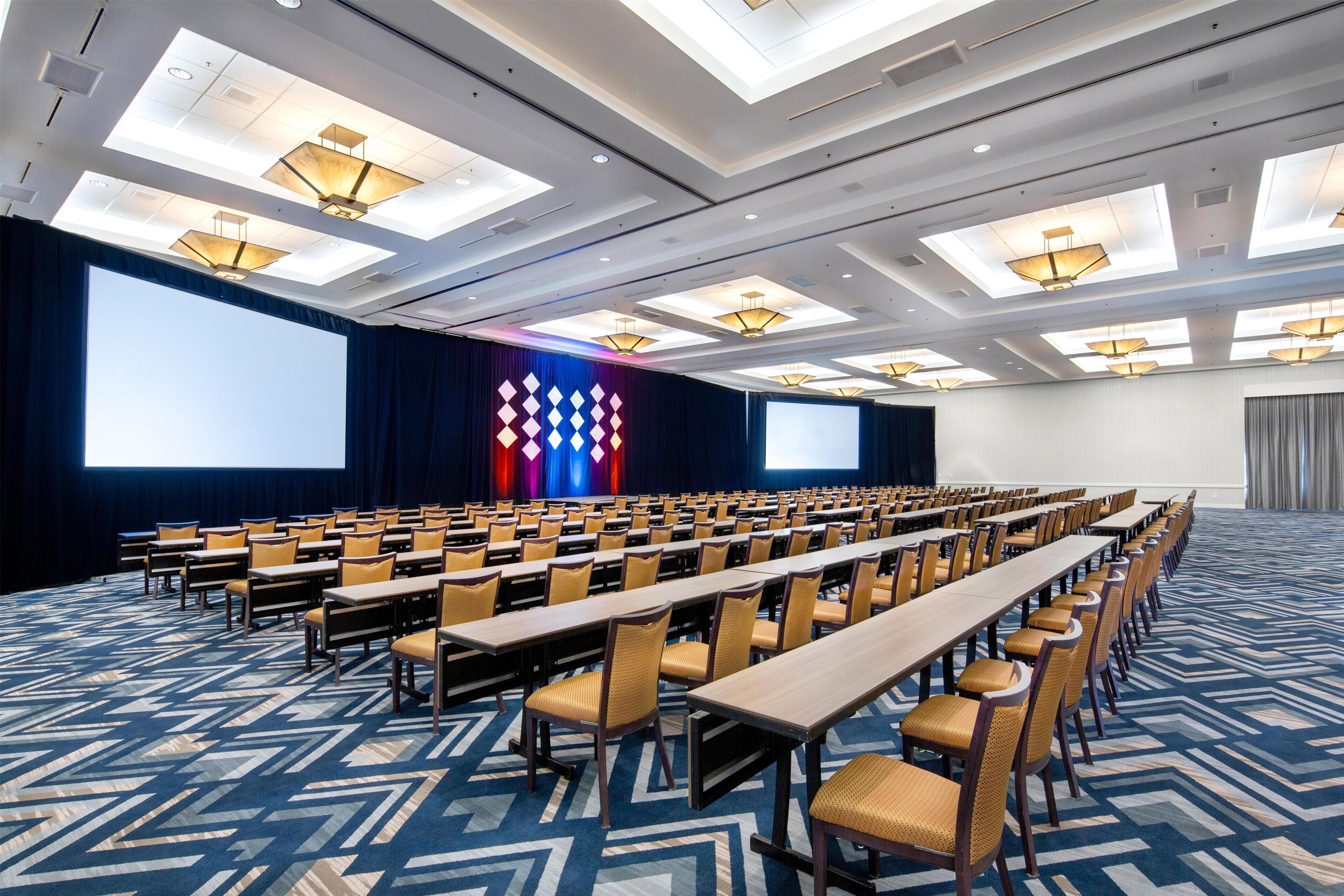 Une salle de conférence avec des rangées de tables et de chaises, un grand écran et des luminaires au plafond.