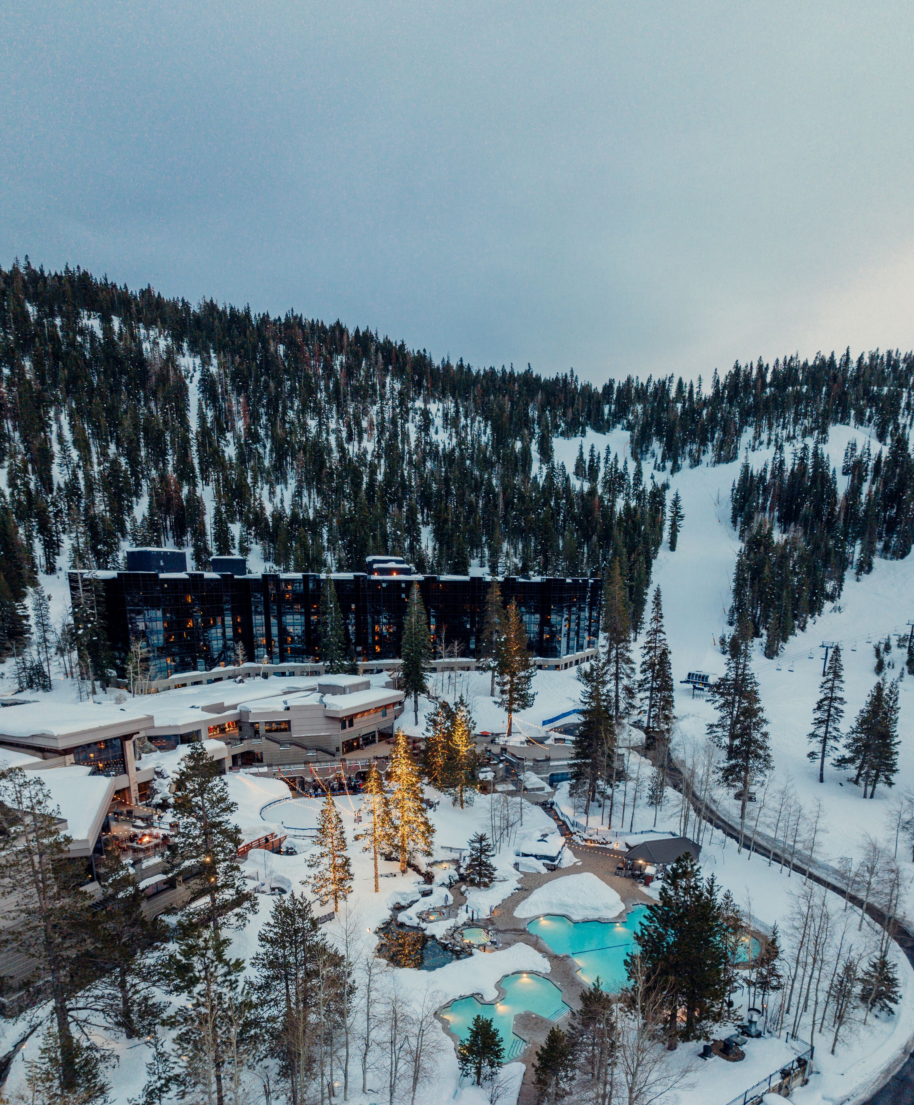 Estância coberta de neve com vários edifícios, piscinas ao ar livre e montanhas ao fundo.