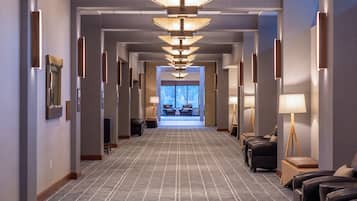 A hotel corridor with a patterned carpet, wall-mounted lamps, and seating areas.