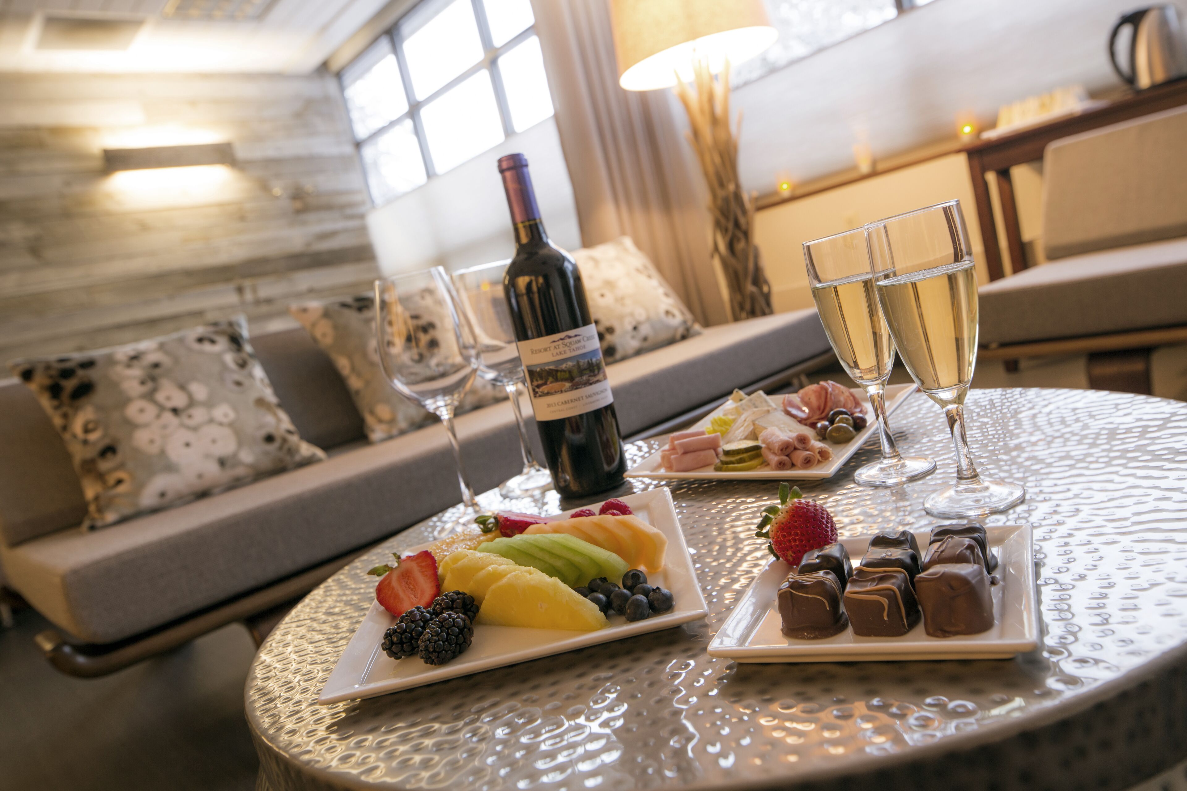 A dining table with a bottle of wine, two glasses of champagne, and plates of fruit and chocolates.