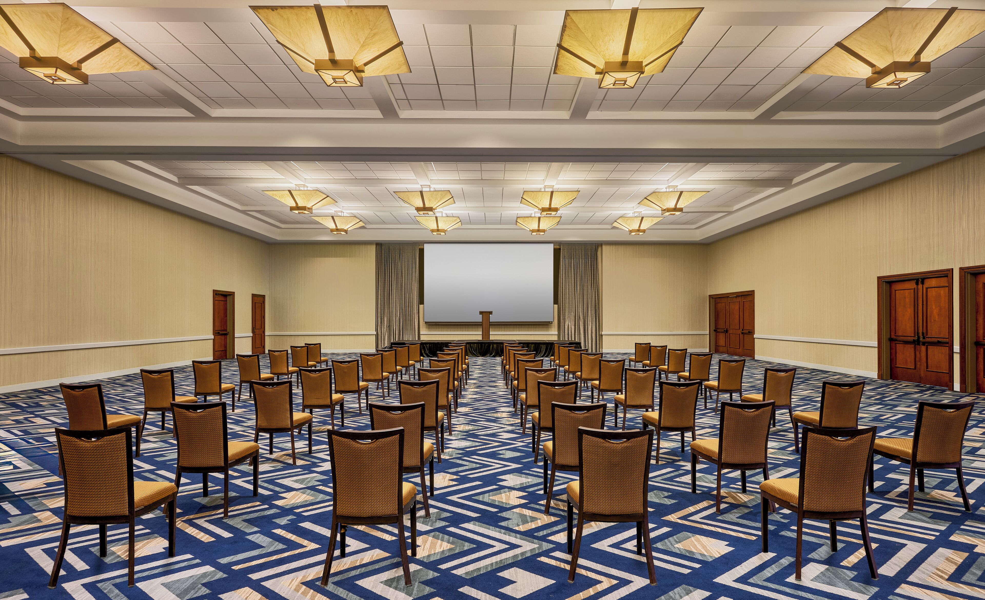 Un salón de conferencias con filas de sillas, una pantalla grande y luces en el techo.