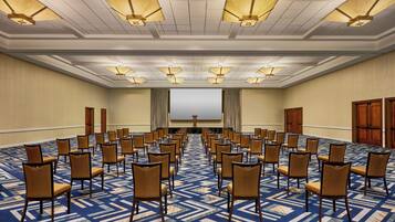 Un salón de conferencias con filas de sillas, una pantalla grande y luces en el techo.