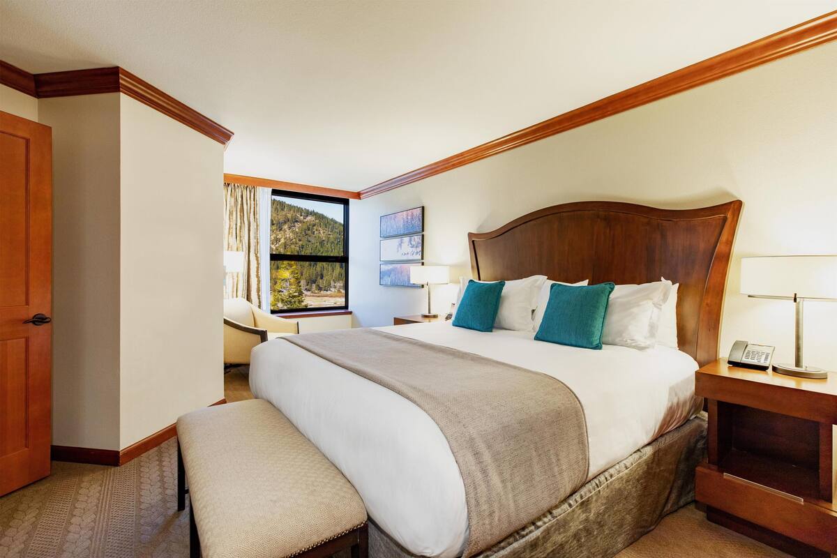 A hotel room with a large bed, a bench, a nightstand with a lamp, a window with a view, and framed pictures on the wall.