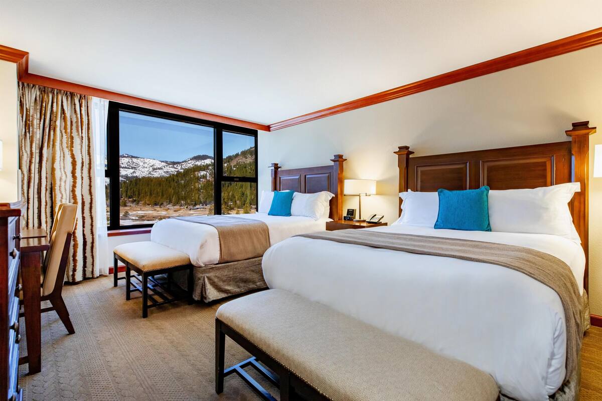 A hotel room with two beds, a desk, a chair, and a large window with a mountain view.