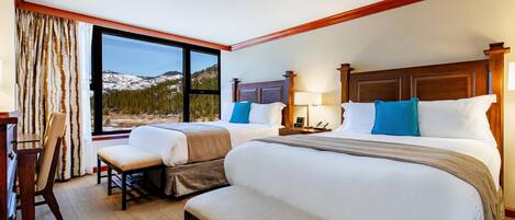 A hotel room with two beds, a desk, a chair, and a large window with a mountain view.