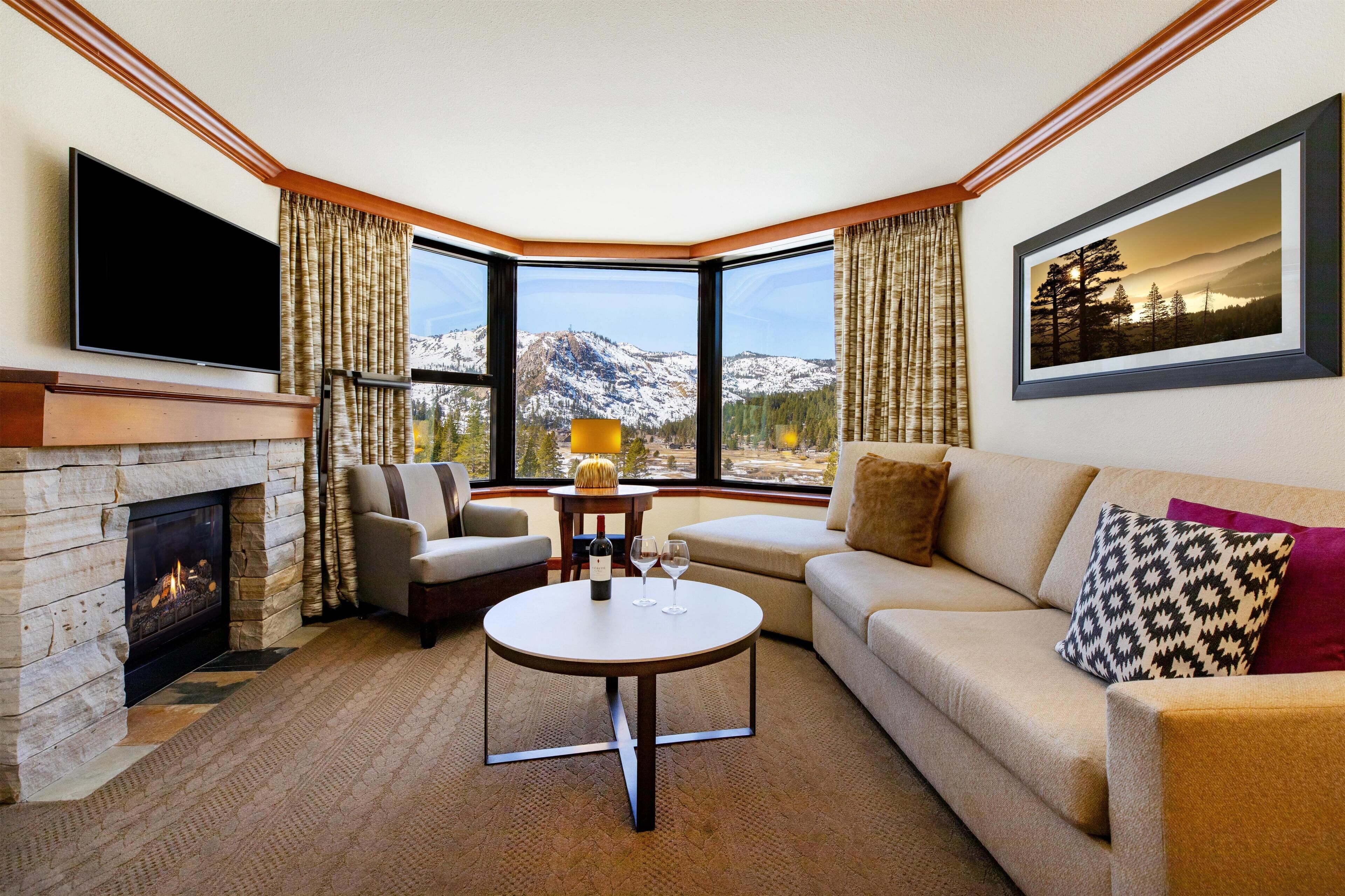 A living room with a fireplace, a television, a sofa, a coffee table, and a large window with a mountain view.